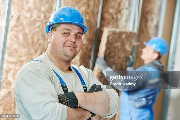 Construction worker in a blue hard hat and overalls standing with arms crossed, insulation installation in the background.