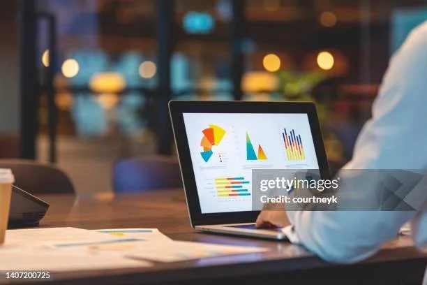 Person analyzing colorful charts on a laptop screen in an office setting with blurred background lights.