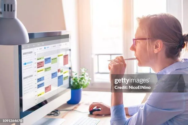 Person using desktop computer with calendar on screen, holding pencil, next to window with natural light, small plant on desk.