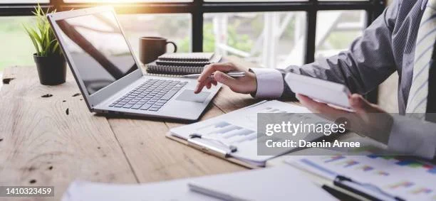 Person using a laptop and holding a smartphone at a wooden desk with documents, pen, coffee cup, and plant.