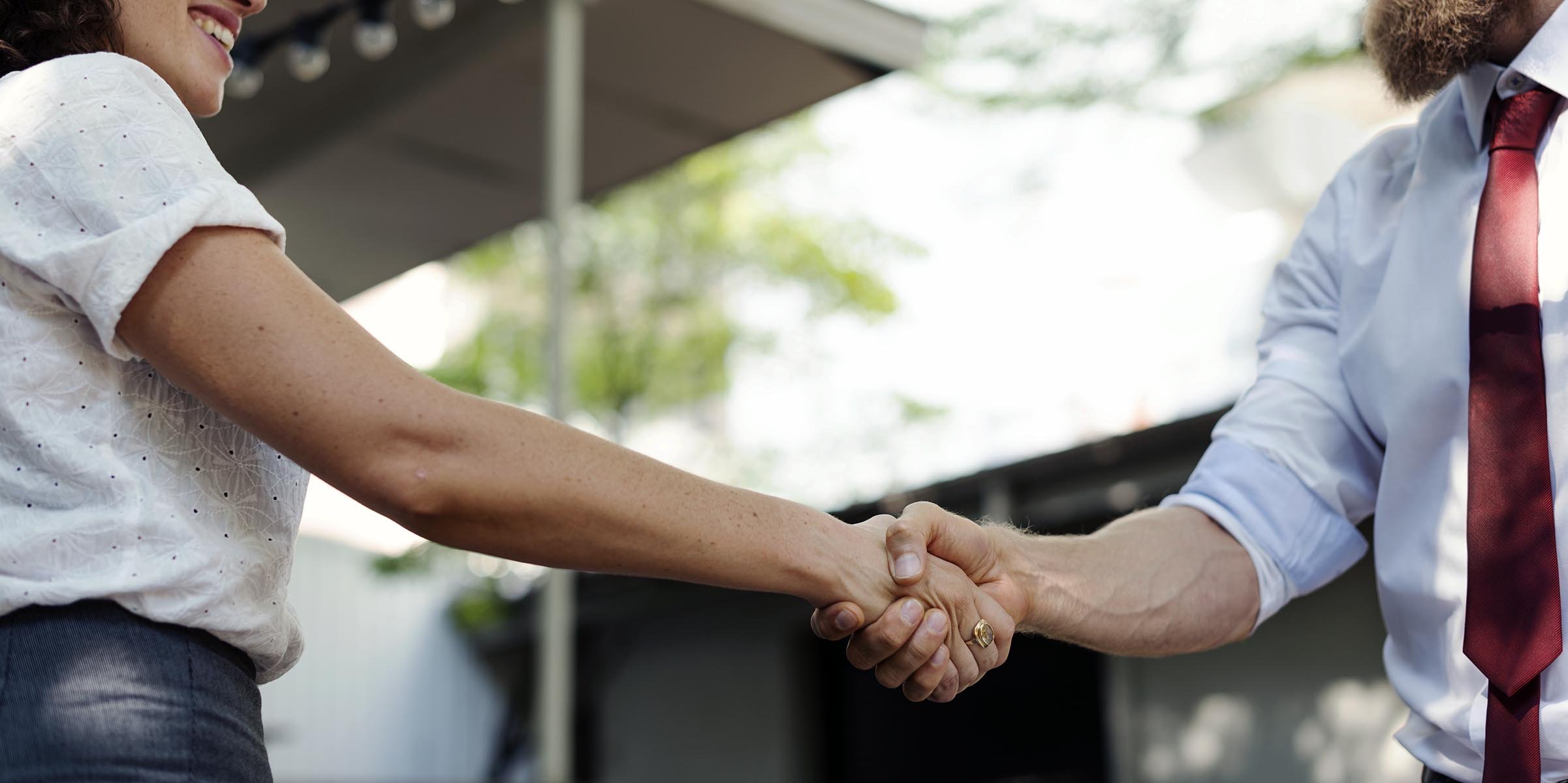 Two people shaking hands outdoors, one wearing a white shirt and the other a blue shirt with a red tie.