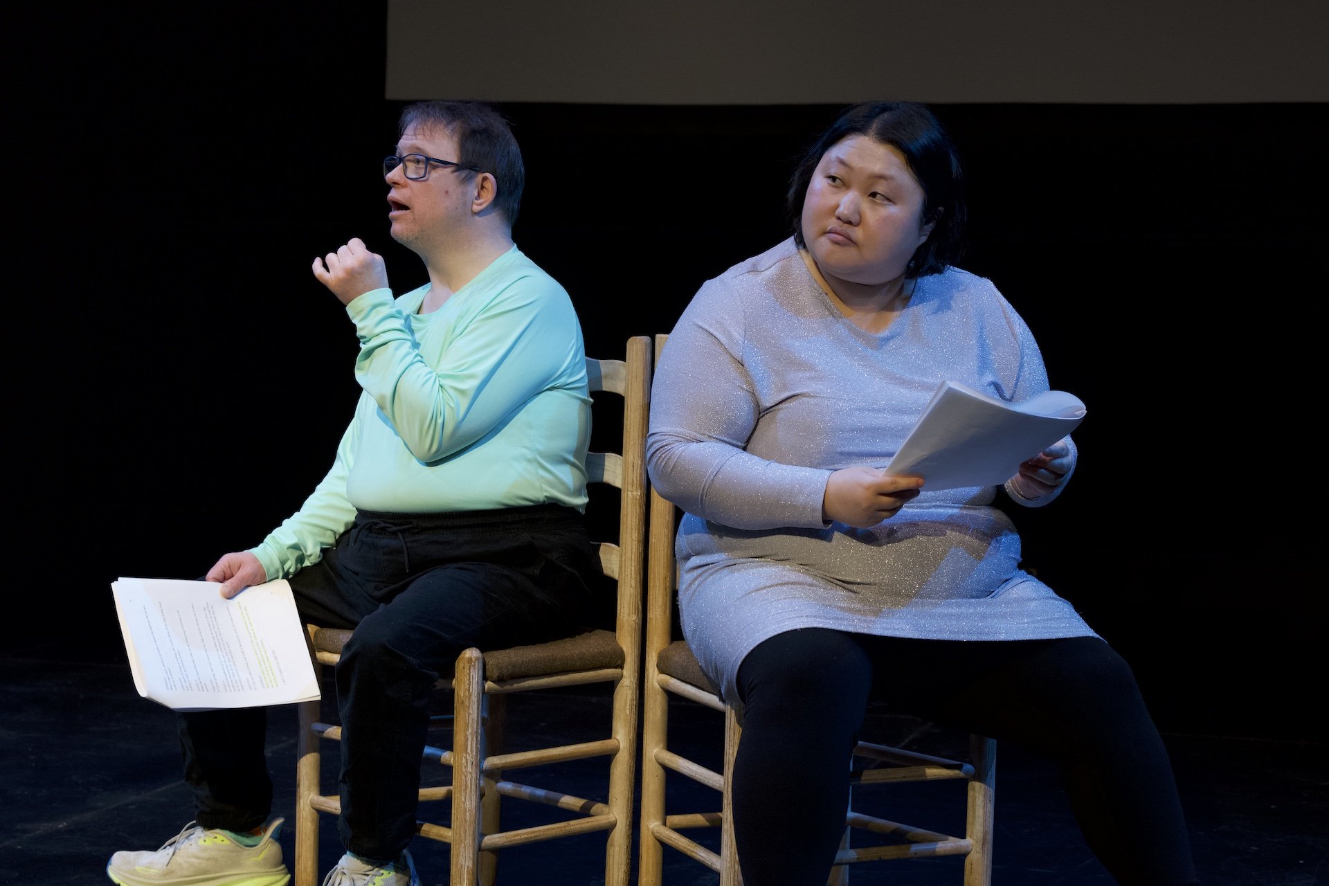 Photo of Andreas Prinz and Angela Sun by Tyler Preece. Andreas and Angela sit on chairs side by side, angled outwards. Andreas wears glasses, a long-sleeved blue shirt, black pants and sneakers. Angela wears a sparkly grey/purple dress, black tights