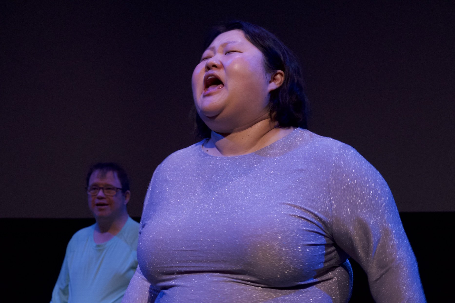 Photo of Angela Sun by Tyler Preece. Andreas Prinz is in the background. Angela wears a sparkly grey/purple dress, black tights and white Converse high tops. She speaking, mouth open.