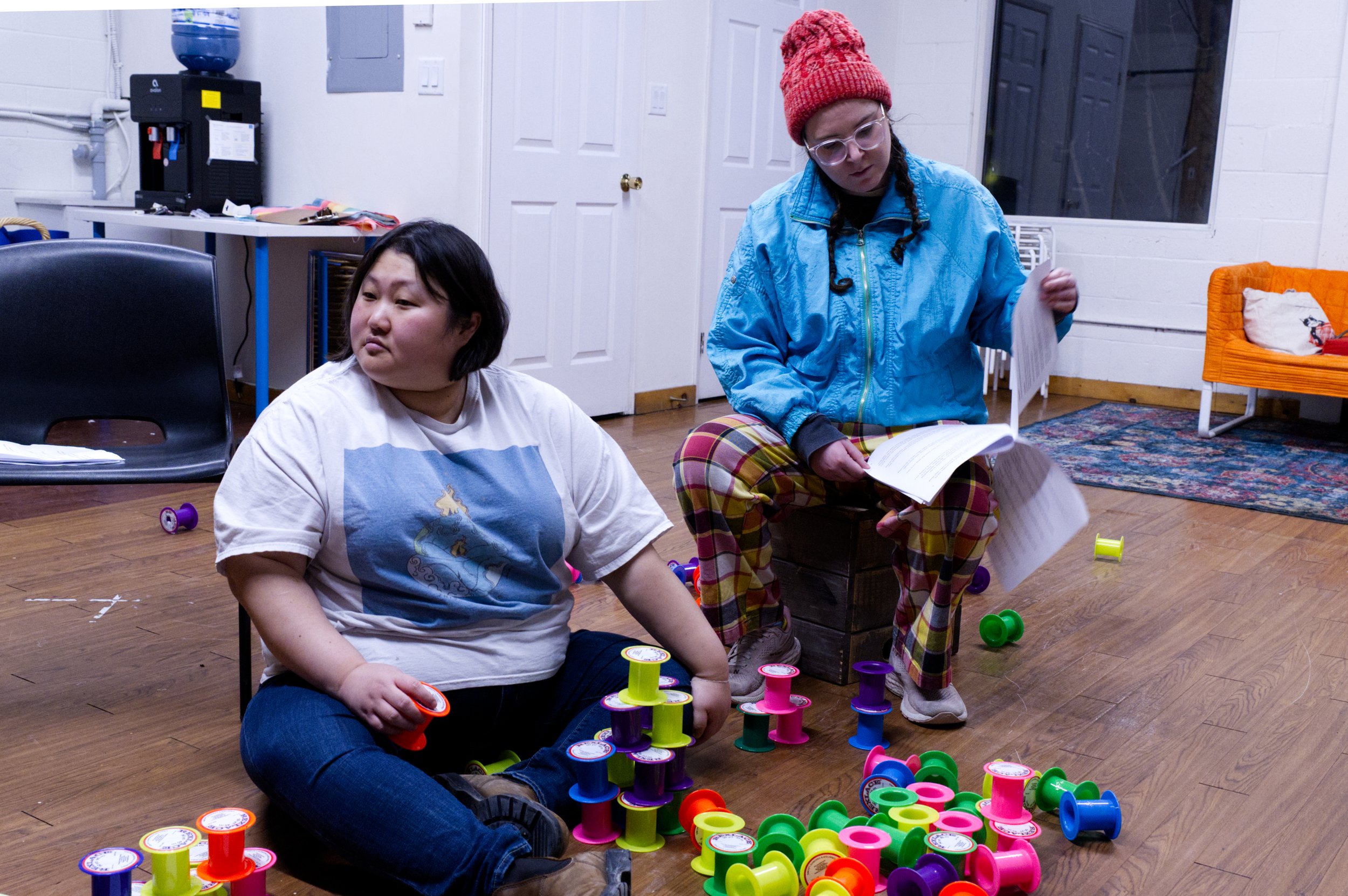 Photo of Angela Sun and Zoe Tomaras by Tyler Preece. Angela sits cross legged on the floor surrounded by brightly coloured empty spools. Zoe sits on a chair nearby, looking down at their script.