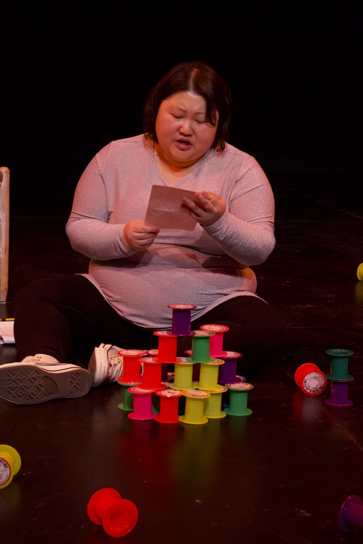 Photo of Angela Sun by Tyler Preece. Angela is sat on the floor, the brightly coloured empty spools stacked in front of her. She reads off of a piece of stationary.