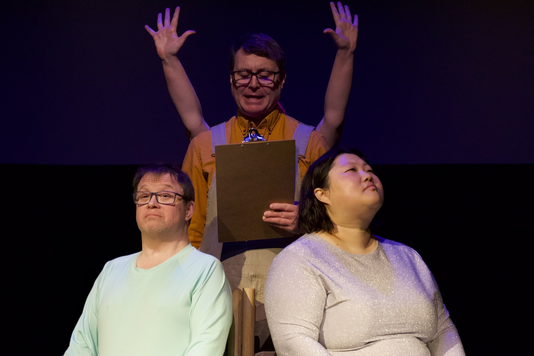 Photo of Andreas Prinz, Timothy Anderson and Angela Sun by Tyler Preece. Andreas and Angela sit on chairs side by side, angled outwards. Timothy stands between them, with Zoe's hands reaching above his shoulders.