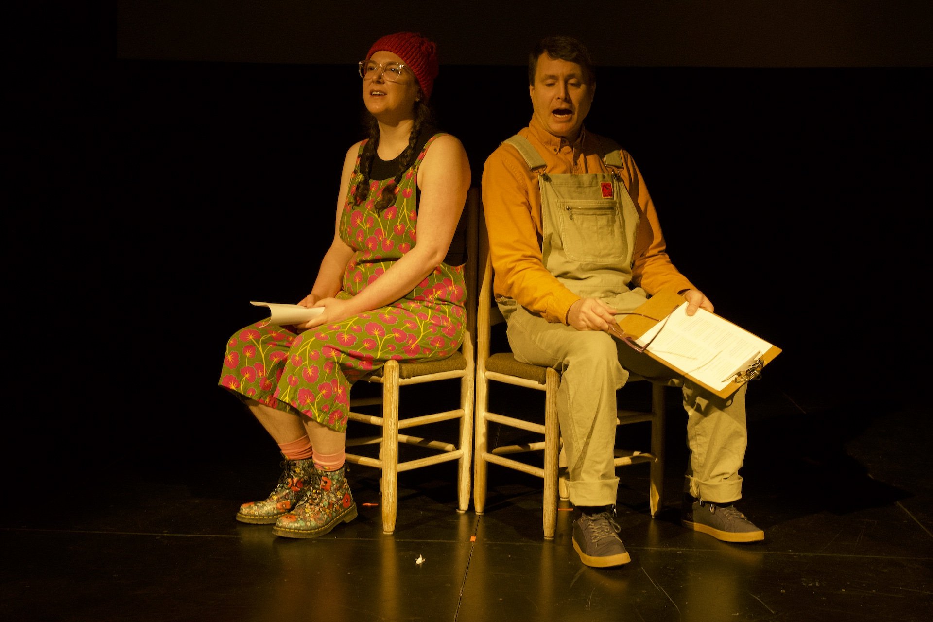 Photo of Zoe Tomaras and Timothy Anderson by Tyler Preece. Zoe and Timothy sit on chairs side by side, angled outwards. Zoe wears clear frame glasses, colourful overalls, floral boots and a red hat. Timothy wears grey overalls, a mustard colour coll