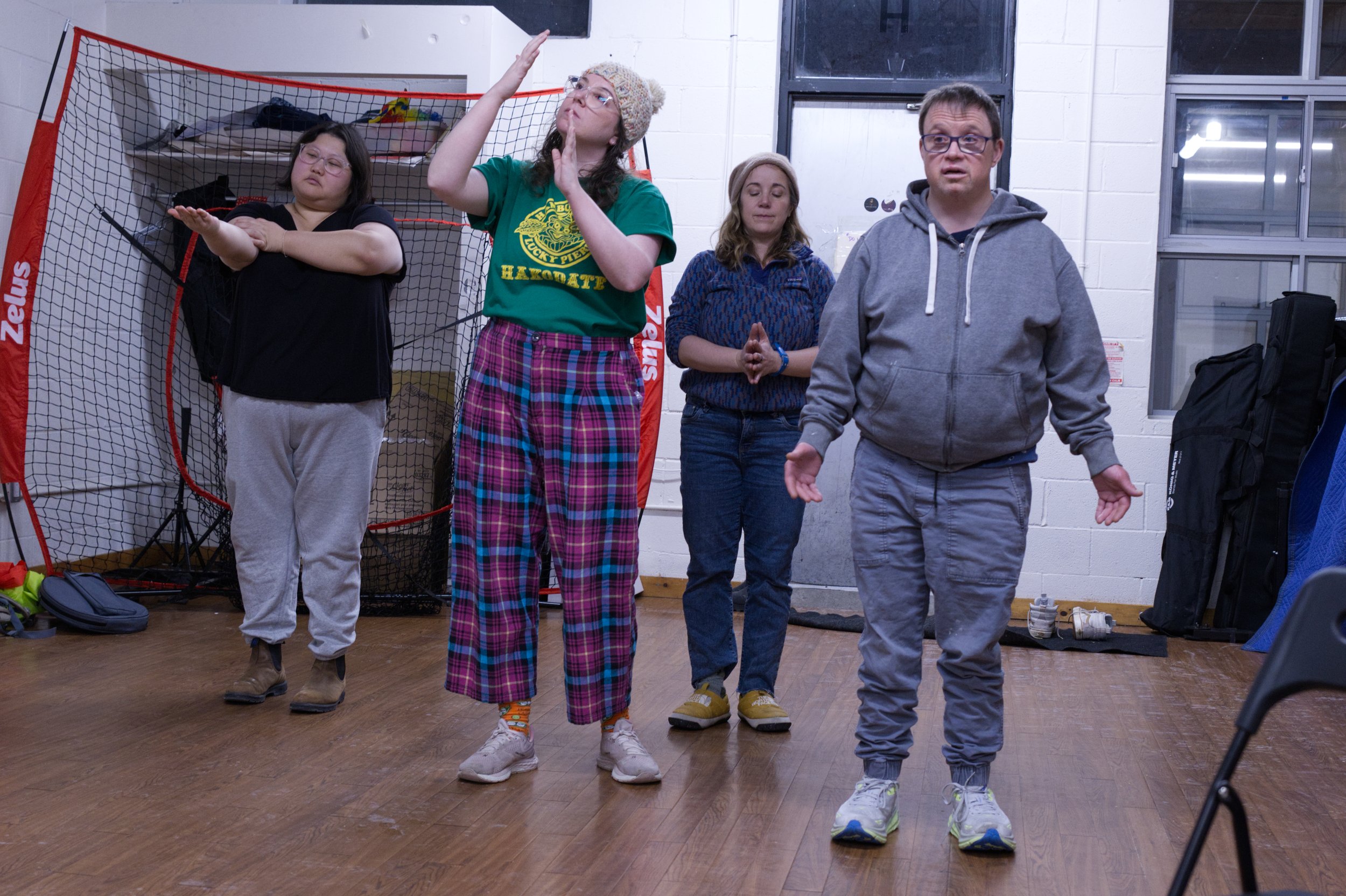 Photo by Tyler Preece. Angela, Zoe, Lisa Marie and Andreas stand in a line, facing the camera. Left to right: Angela stands, bending her right arm in front of her outstretched left arm. Zoe's arms are raised, with their elbow's bent. Lisa Marie's han
