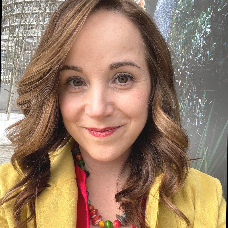 Lisa Marie is a white woman with light brown hair, wearing a colourful necklace, red shirt and yellow jacket.