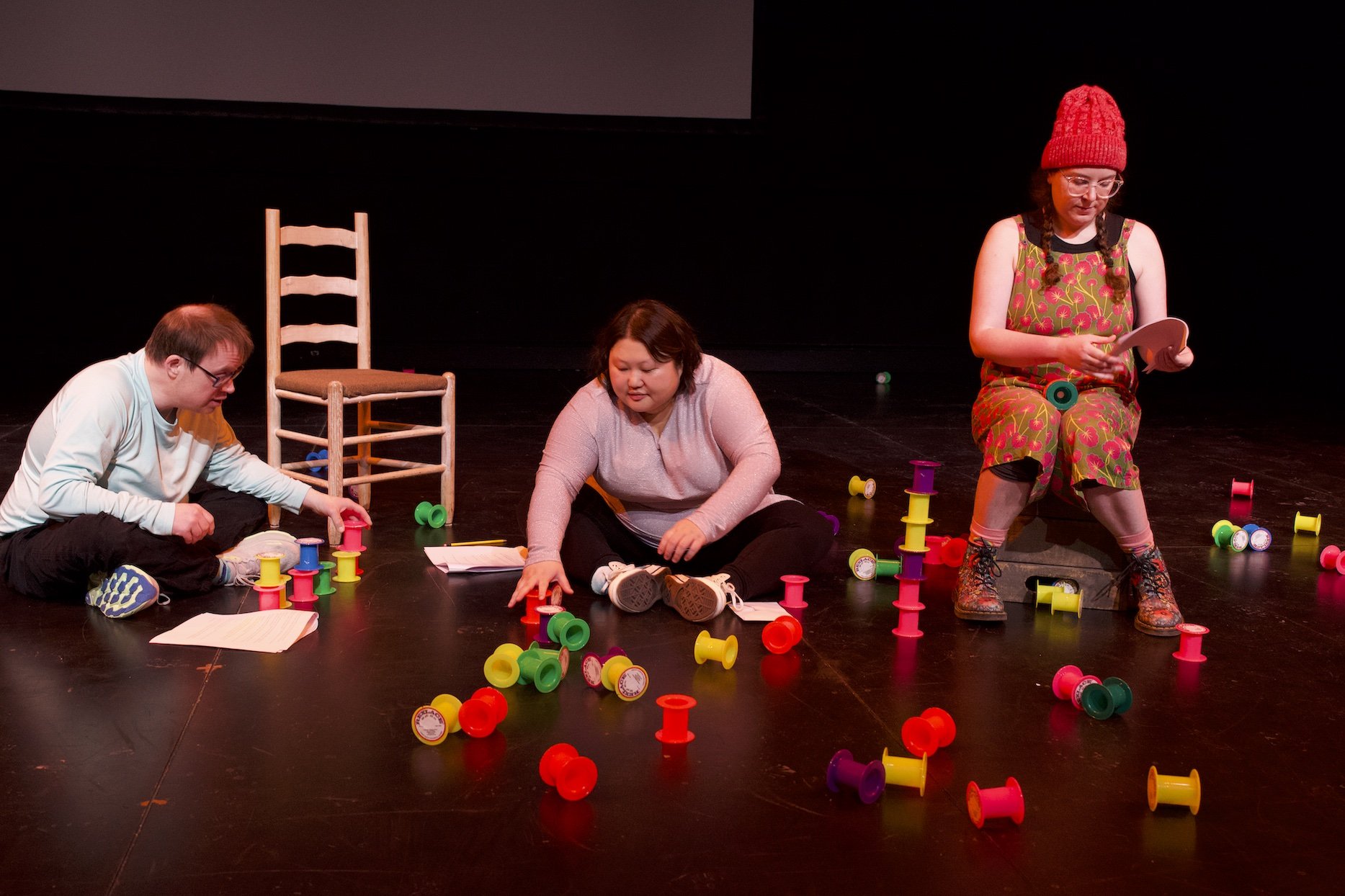 Photo by Tyler Preece. From left, Andreas Prinz, Angela Sun and Zoe Tomaras. Andreas and Angela are sat on the floor, amongst brightly coloured empty spools. Zoe is sat on a chair.