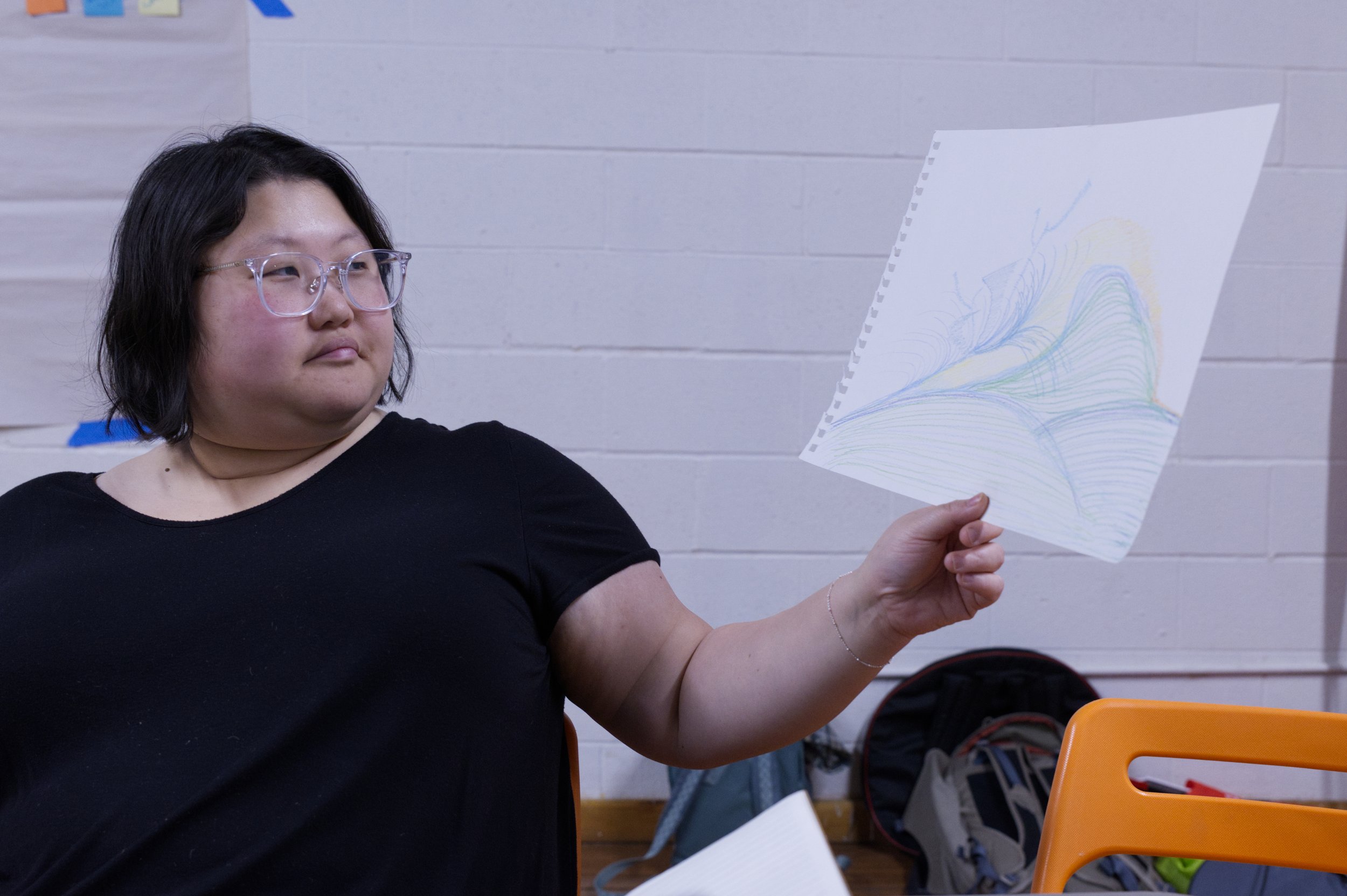 Photo of Angela Sun by Tyler Preece. Angela wears clear framed glasses and a black t-shirt. She holds up a drawing.