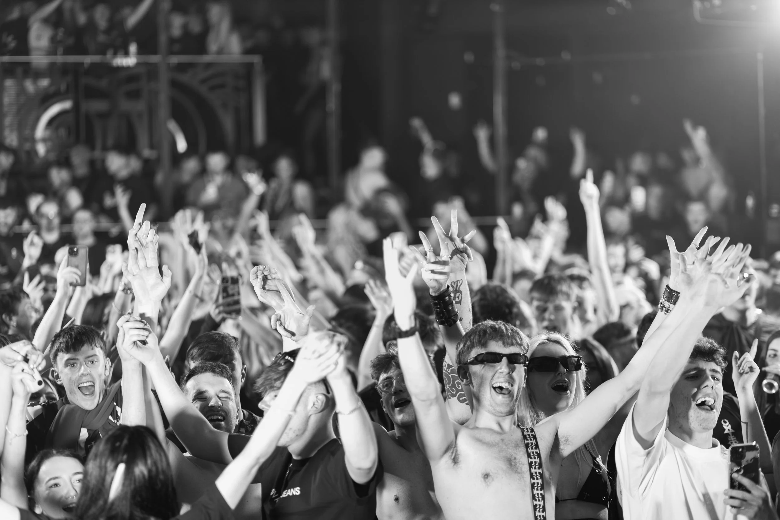 Crowd of people at a concert or event, many with hands raised, taking photos, and wearing sunglasses, in a lively atmosphere.