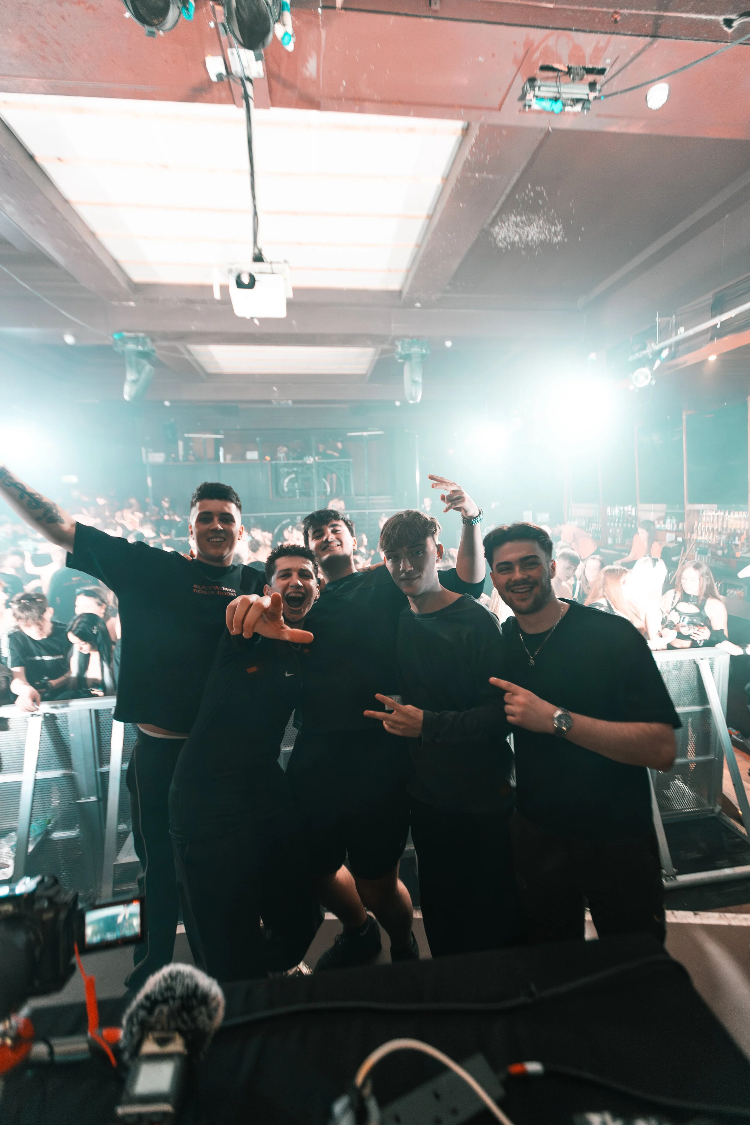 Five young men posing together at a concert, smiling and making gestures in a lively nightclub with bright stage lights, crowd, and performance equipment in the background.