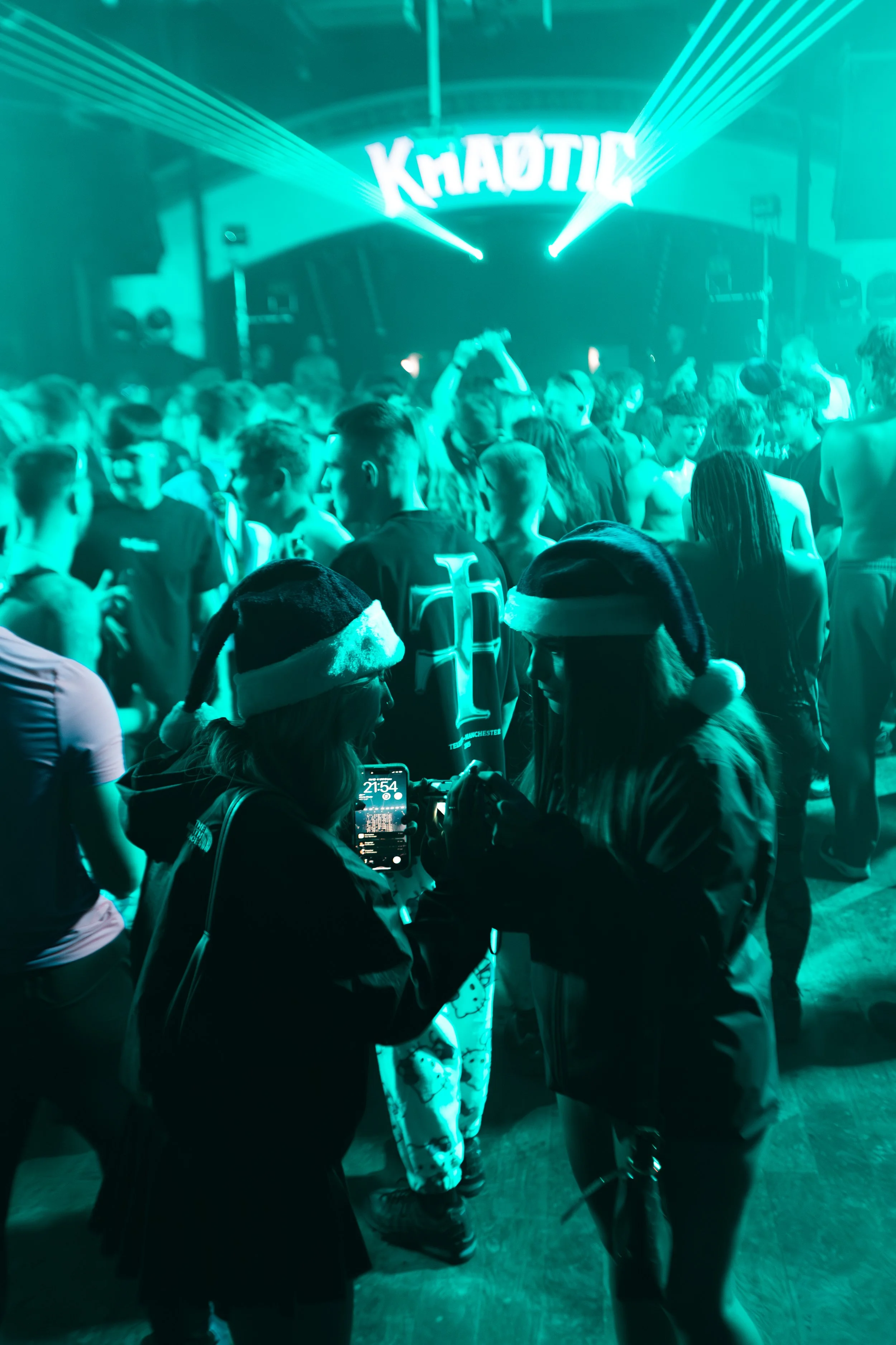 People at a holiday event wearing Santa hats, looking at mobile phones, with a crowded dance floor and a sign that reads 'KHAOTIC' in the background, illuminated by blue-green lighting.