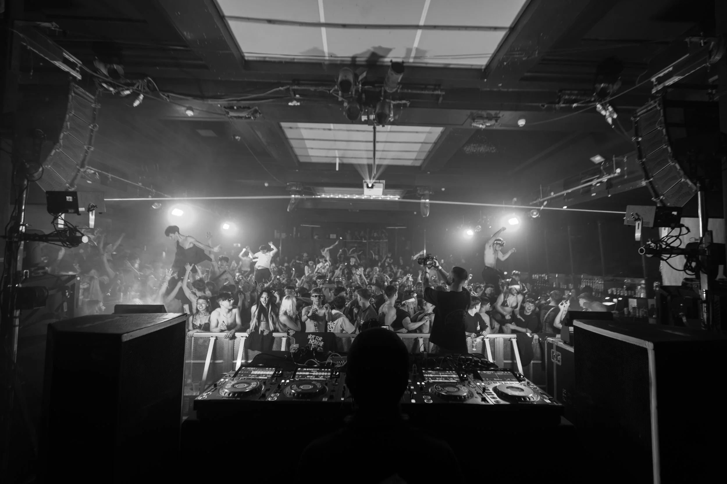 Black and white photo of a crowded dance club with a DJ at the front, a large group of people dancing, and bright lights illuminating the scene.