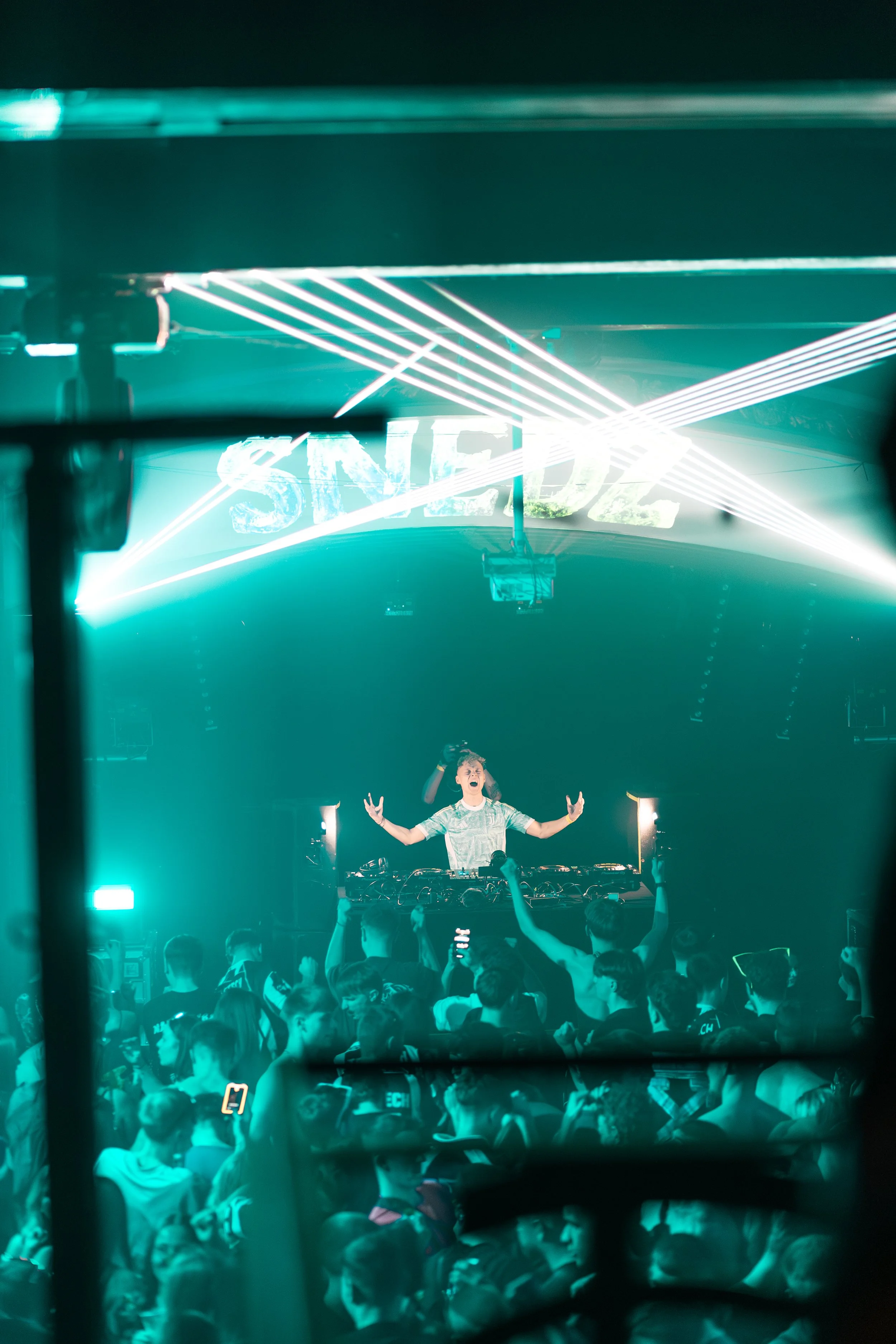A DJ performs on stage at a concert with laser lights, surrounded by a crowd of fans, some holding up phones, under a sign that spells 'SNEE' in bright lights.