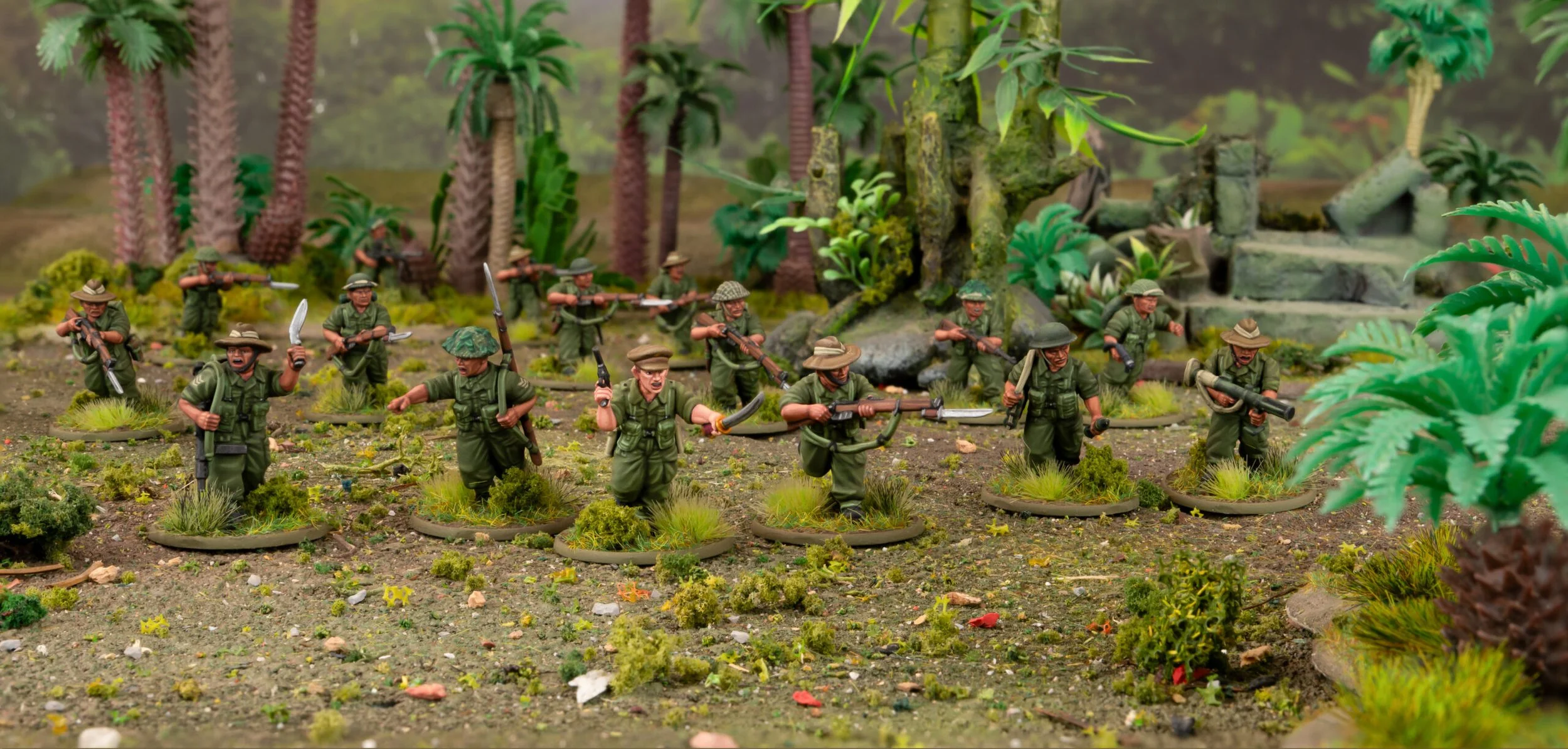 Miniature soldiers in green uniforms and hats, armed with rifles and weapons, are positioned on a terrain with scattered grass and small plants, surrounded by artificial trees and jungle-like scenery.