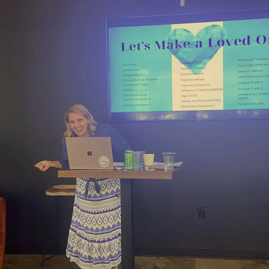 A woman with blonde hair, wearing a blue jacket over a white and blue patterned skirt, smiling and standing at a high table with a laptop, snacks, and drinks. Behind her, a screen displays a presentation titled "Let's Make a Loved O" with a large blue heart at the top.