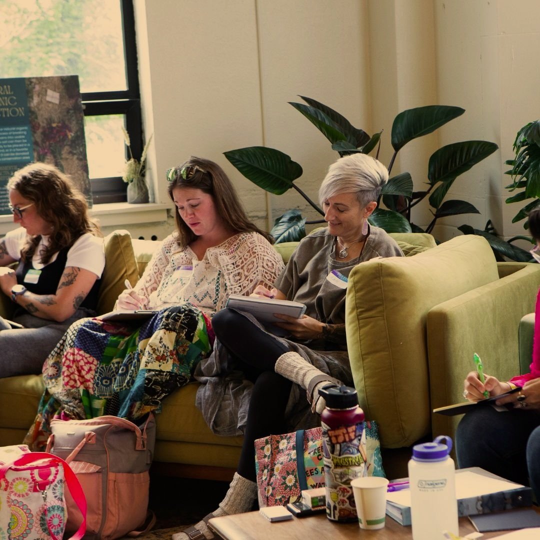 A group of people sitting on a yellow couch taking notes, surrounded by plants and personal items such as bags and cups in a cozy indoor setting.