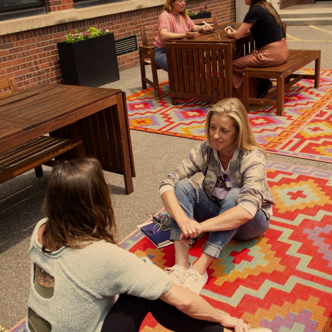 People sitting on colorful rugs outdoors, talking near wooden tables.