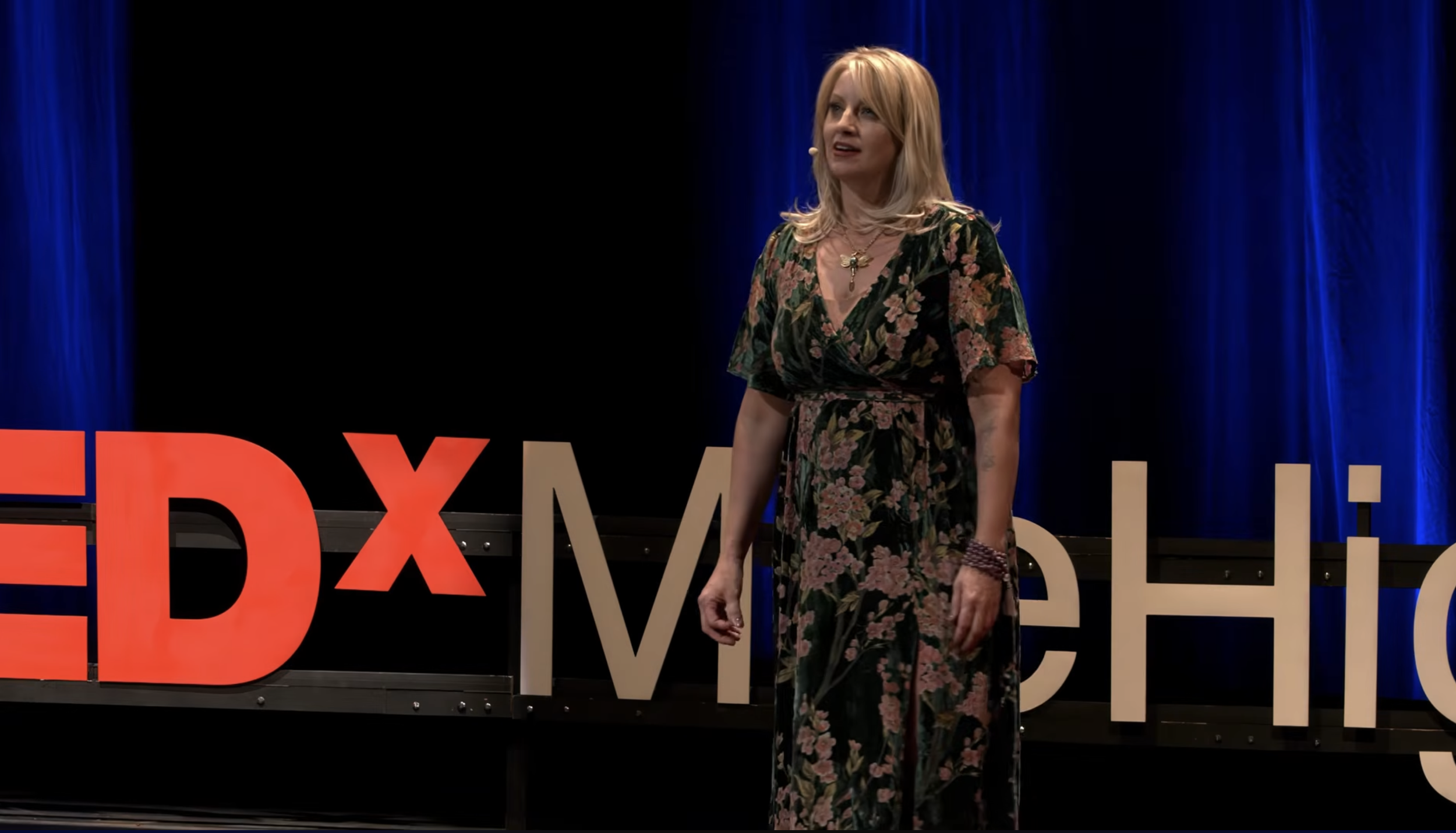 A woman with blonde hair wearing a floral dress standing on a stage with a TEDx sign behind her.