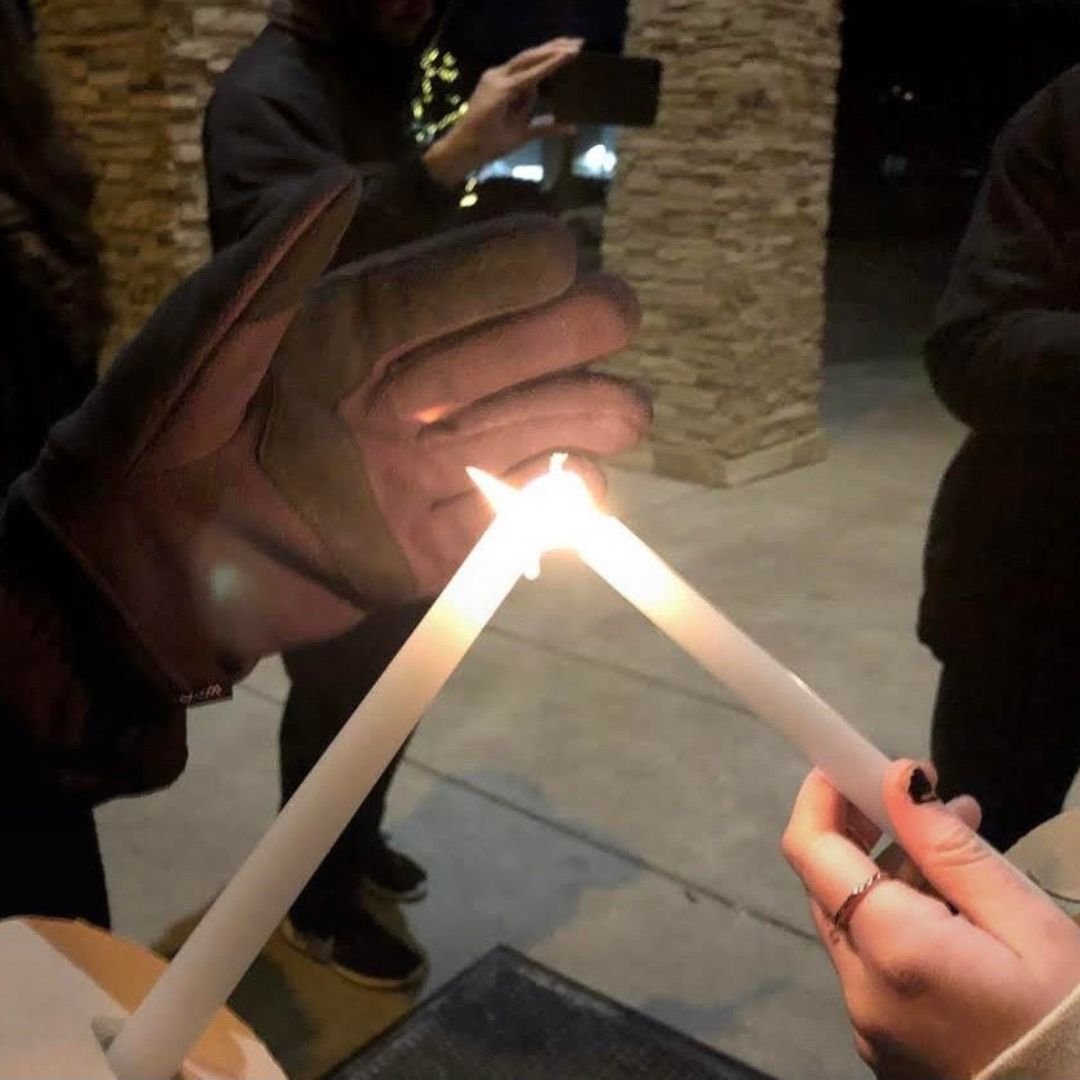 People lighting candles outdoors at night, one person wearing gloves.
