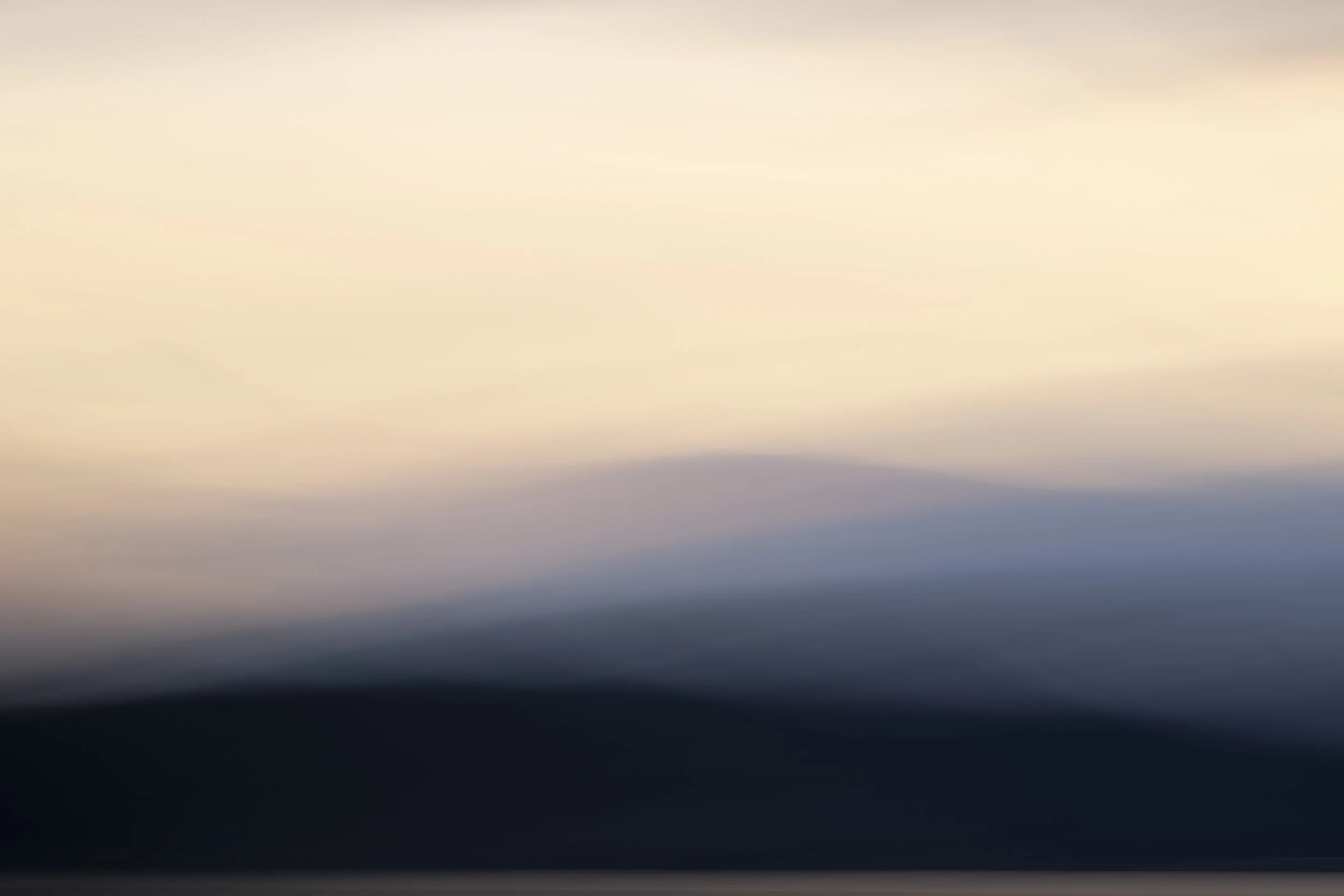 Blurred landscape image with gradient colors of the sky at sunset or sunrise, showing hints of black mountains or hills in the foreground.