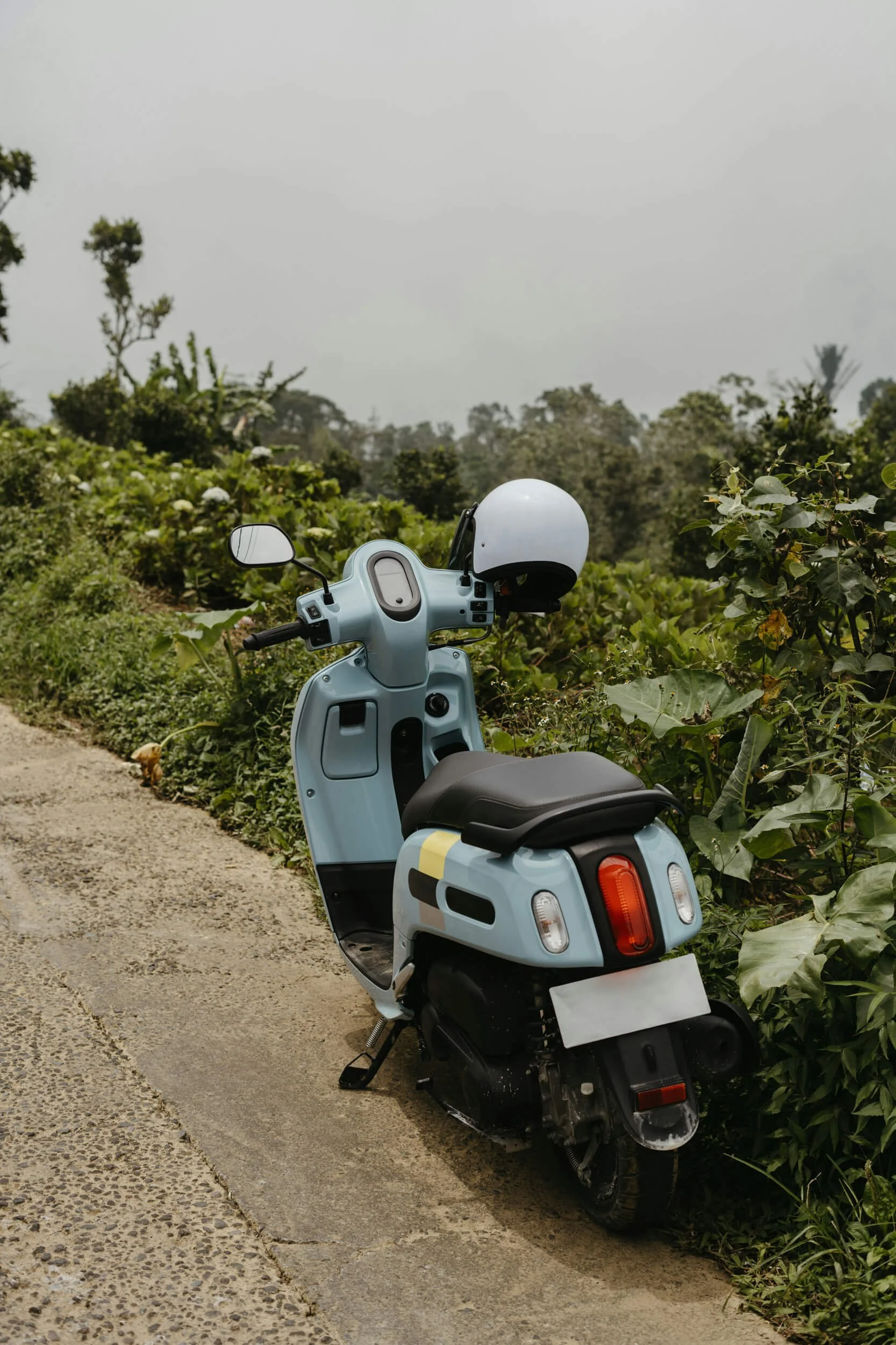 Blue scooter parked on a roadside with greenery and a cloudy sky.