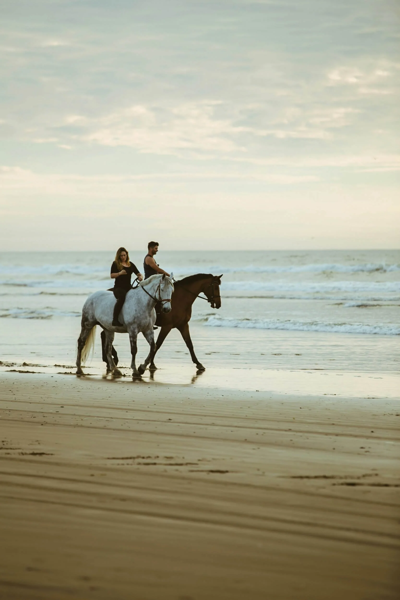 Two people horseback riding on a beach, with the ocean in the background.
