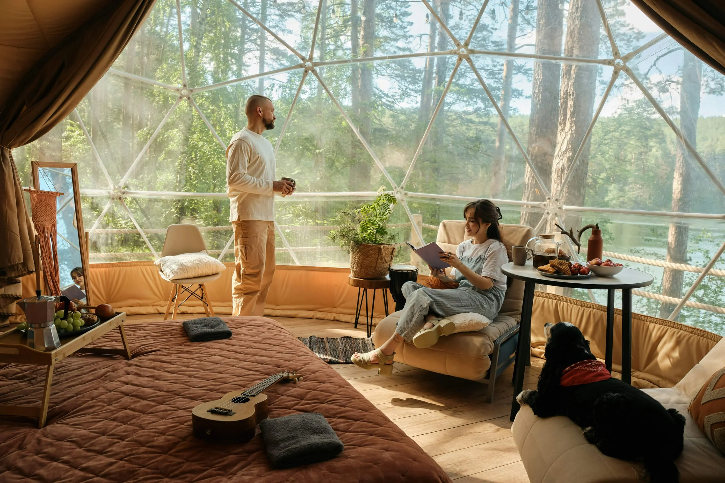 Couple in a geodesic dome with forest view, enjoying leisure time with a dog, food, and ukulele.