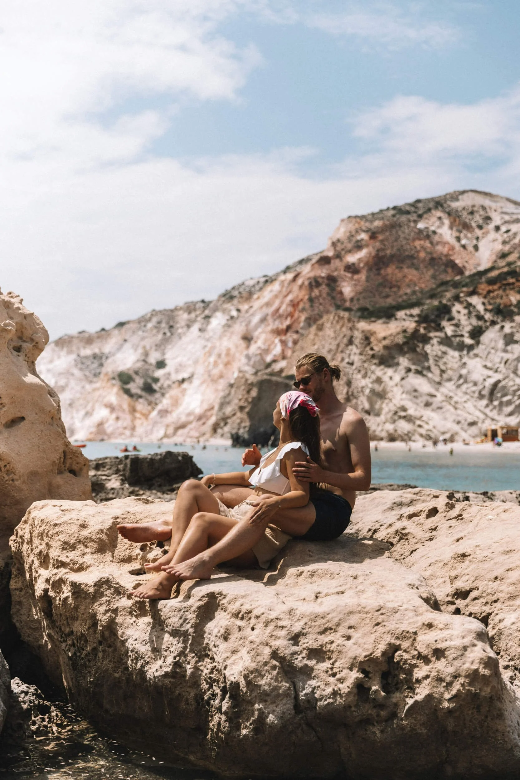 Couple sitting on rocks by the sea with cliffs in the background.