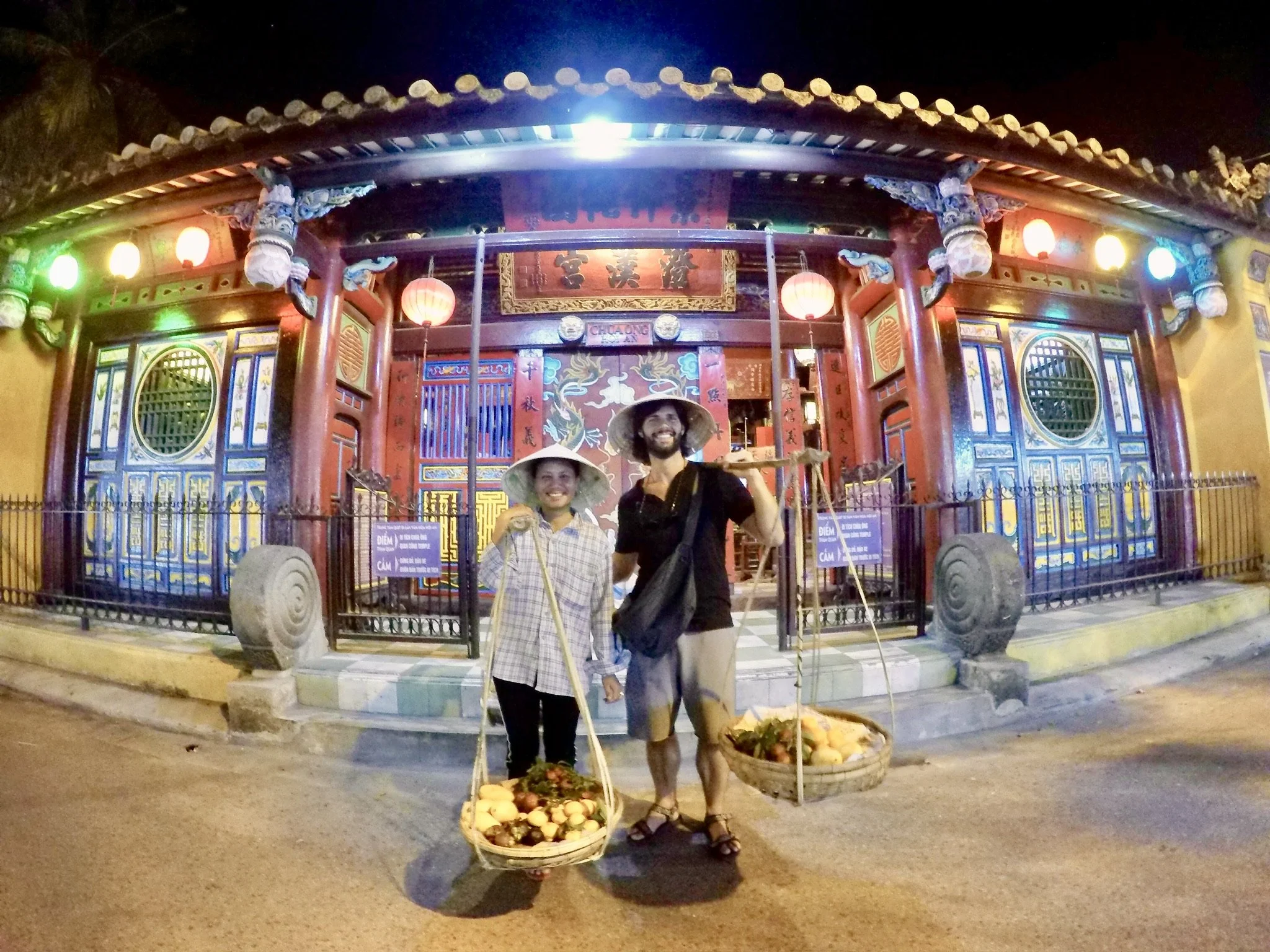 Two people wearing traditional hats and holding fruit baskets in front of a colorful Chinese temple at night, decorated with lanterns.