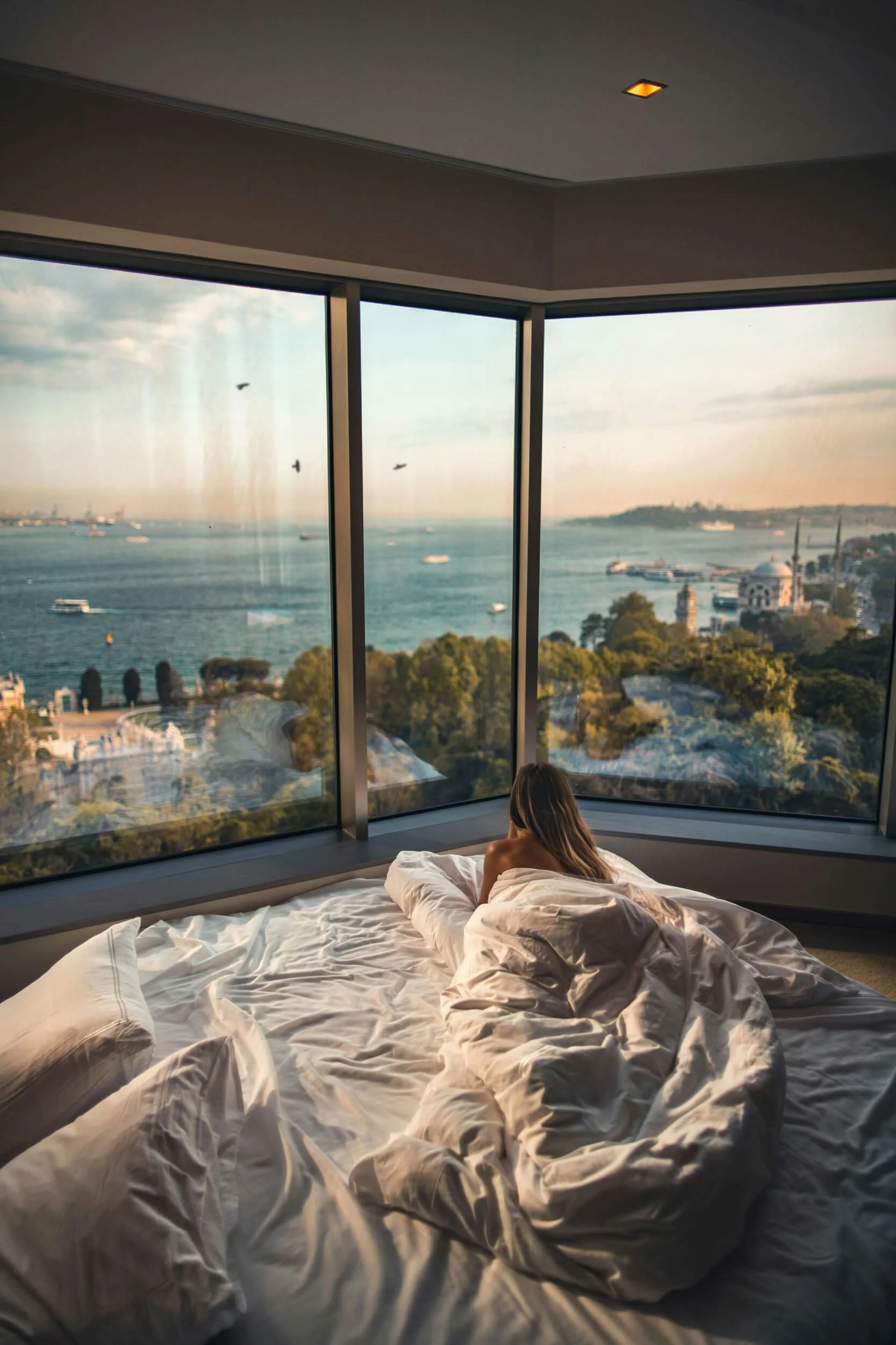 Person lying on a bed with white sheets, looking out a large window at a scenic cityscape and ocean view.