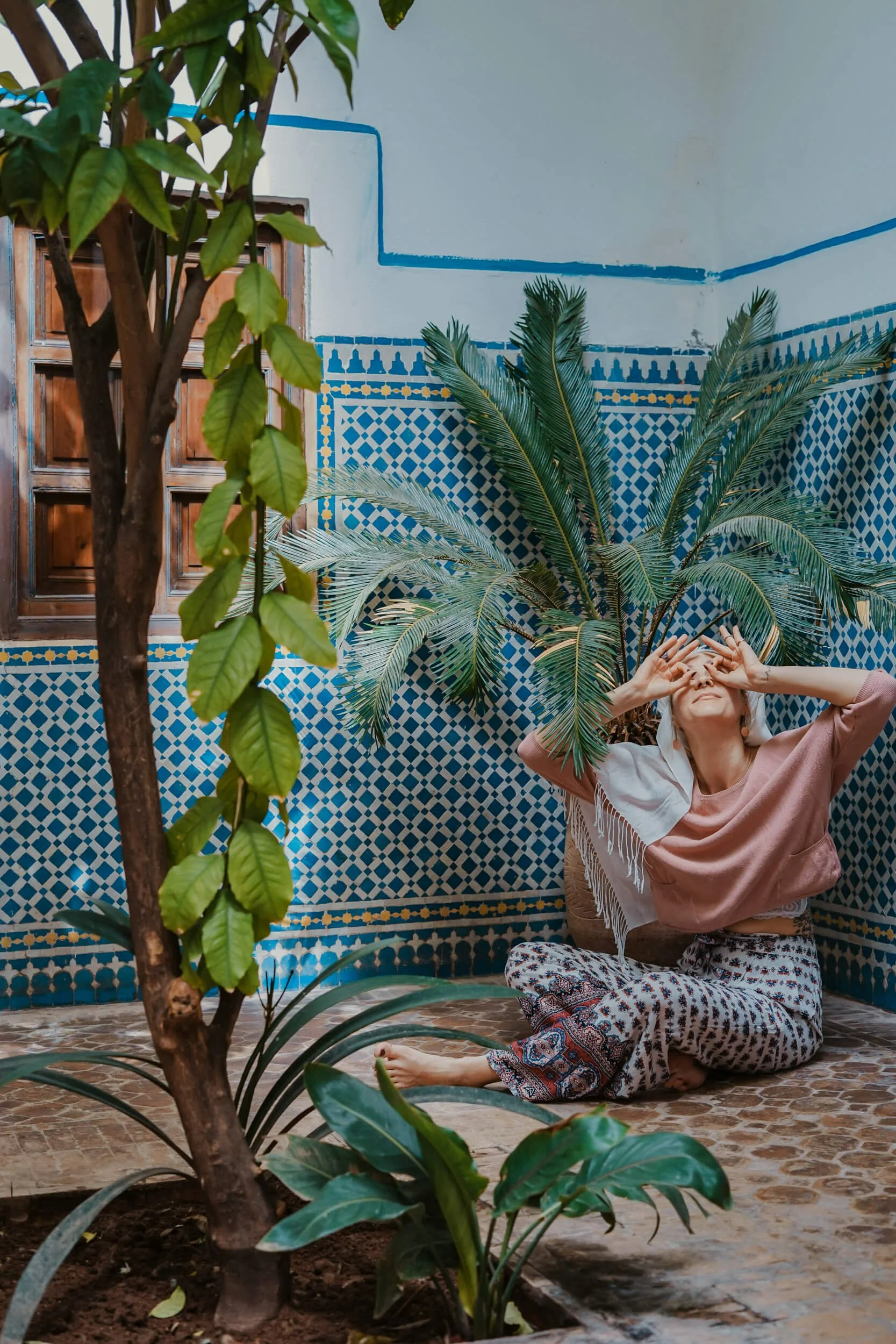 Woman sitting on tiled floor in courtyard with plants, wearing pink top and patterned pants, hands over eyes, near blue and white mosaic wall.