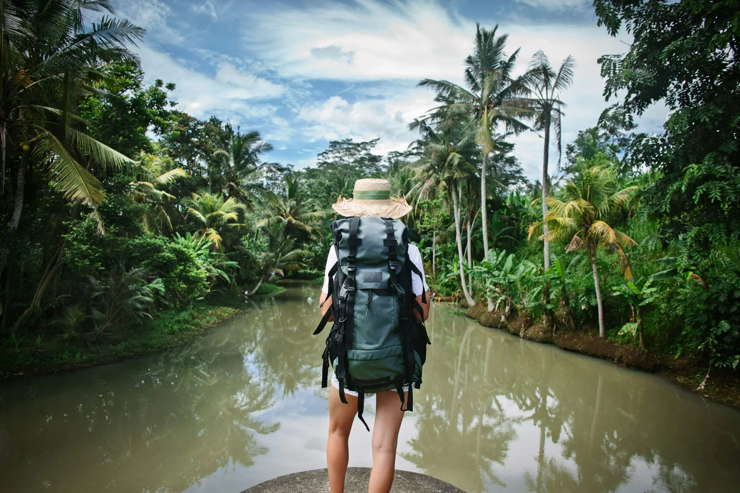 A person wearing a straw hat and large backpack standing by a river surrounded by tropical trees, looking at the lush greenery.