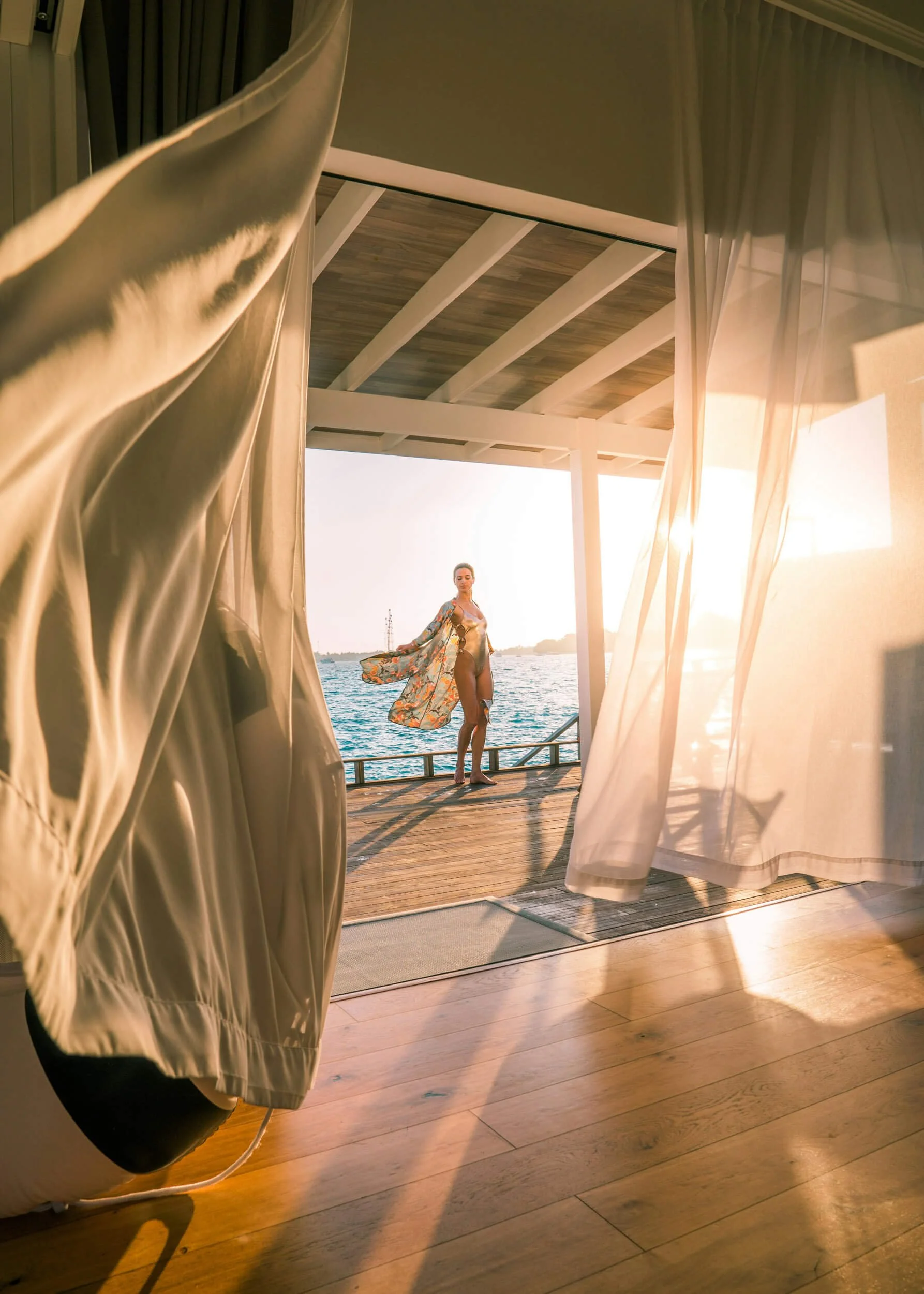 Person standing on a deck by the ocean, wearing a floral robe, with sheer curtains blowing in the foreground and sunlight streaming through.