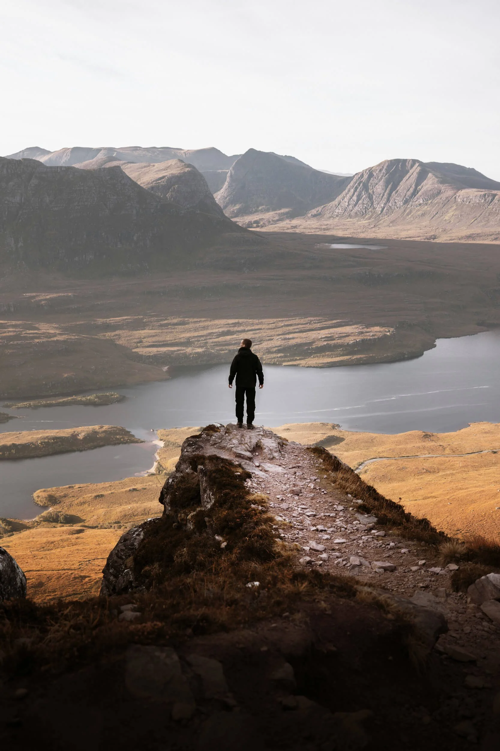 Person standing on a rocky edge overlooking a lake with mountainous landscape.