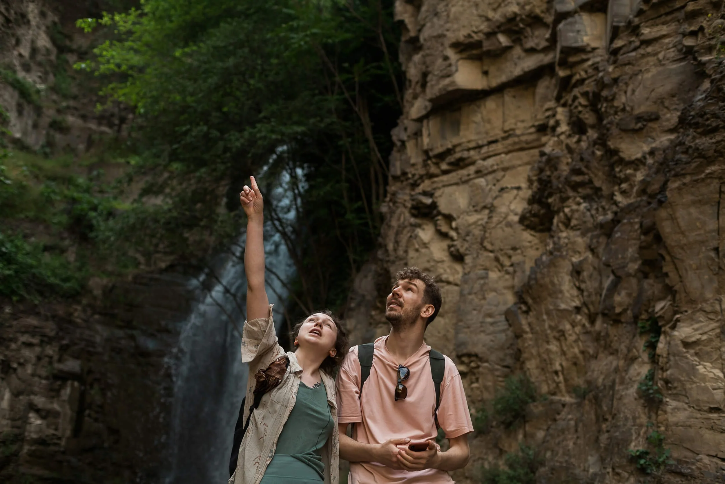Two people hiking near a waterfall and rocky cliff, one pointing upwards.