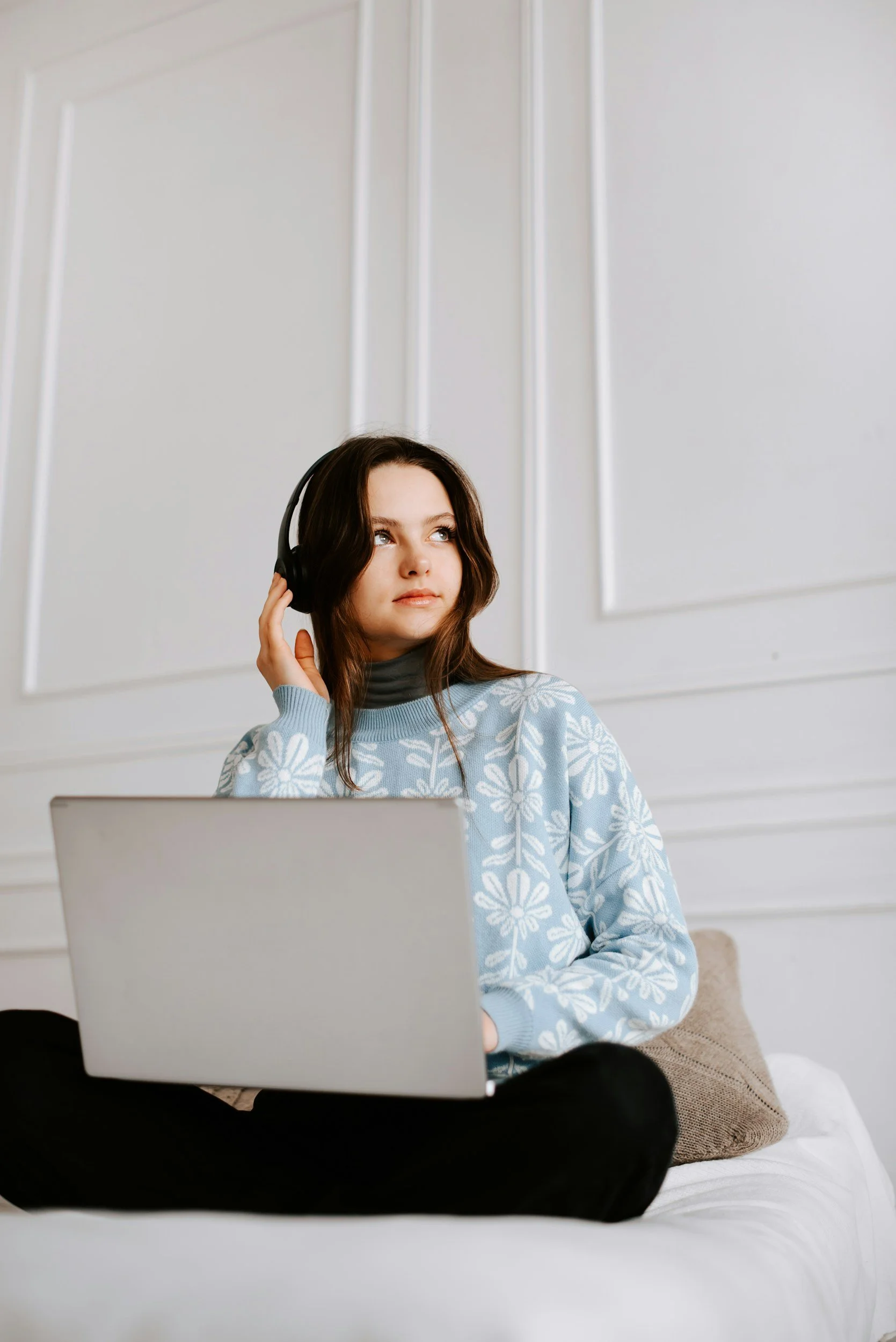A young woman with brown hair wearing a blue sweater with white floral patterns, sitting on a bed with a beige pillow and using headphones while working on a silver laptop.