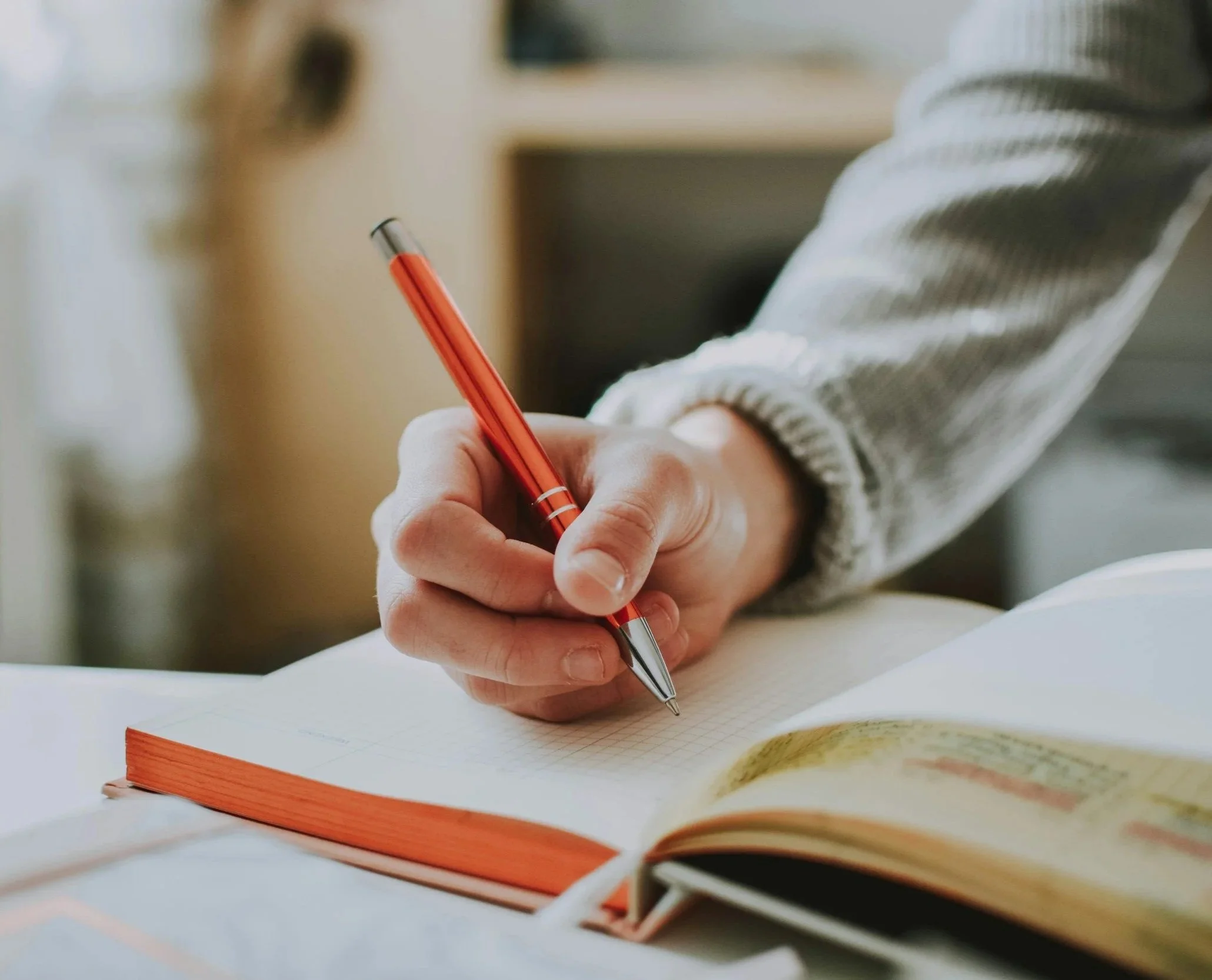 A person wearing a gray and white striped sweater writes in a notebook with a red pen on a white desk.