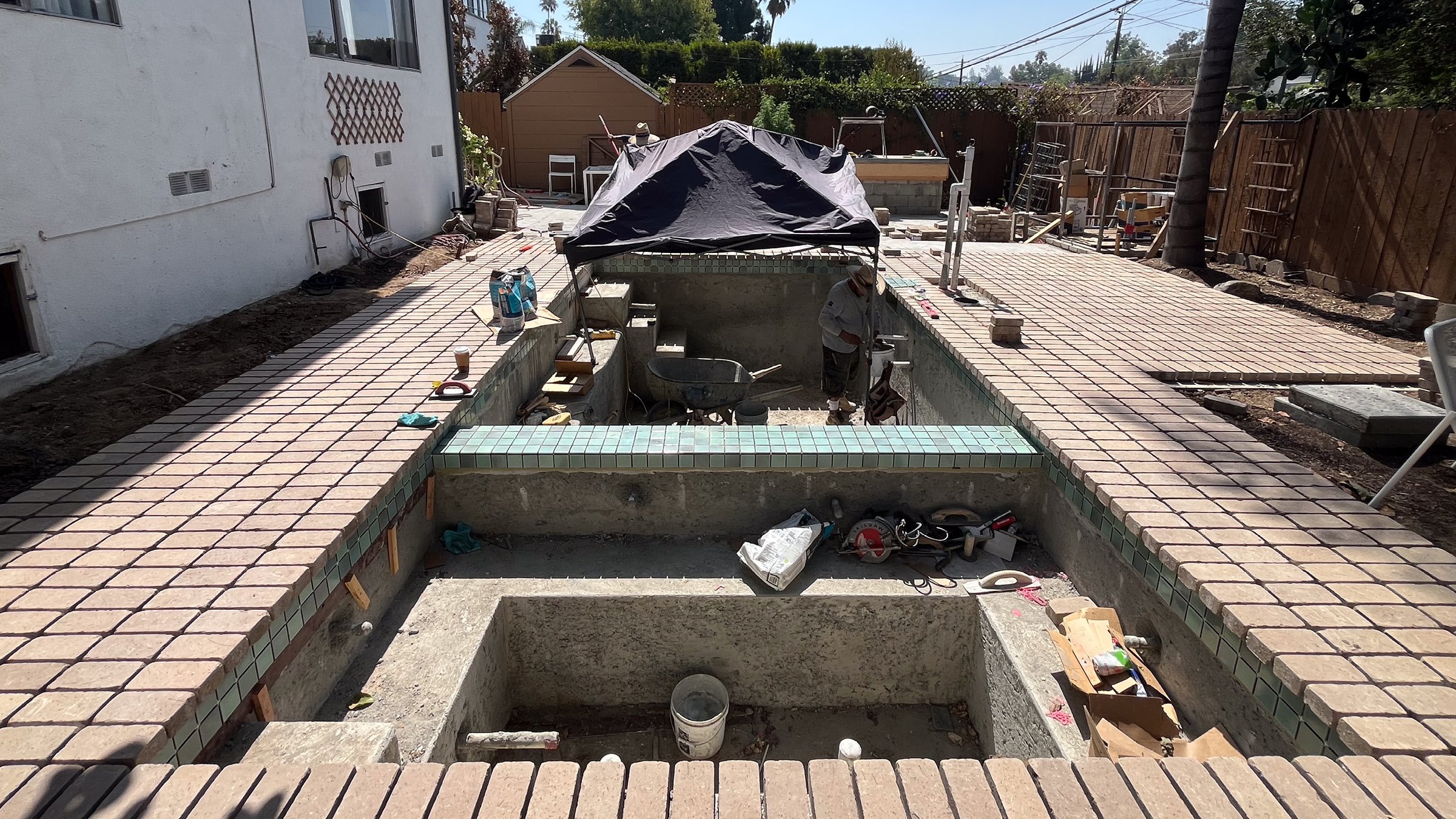 Worker installing brick paving and pale green tile around a new swimming pool and spa under construction.