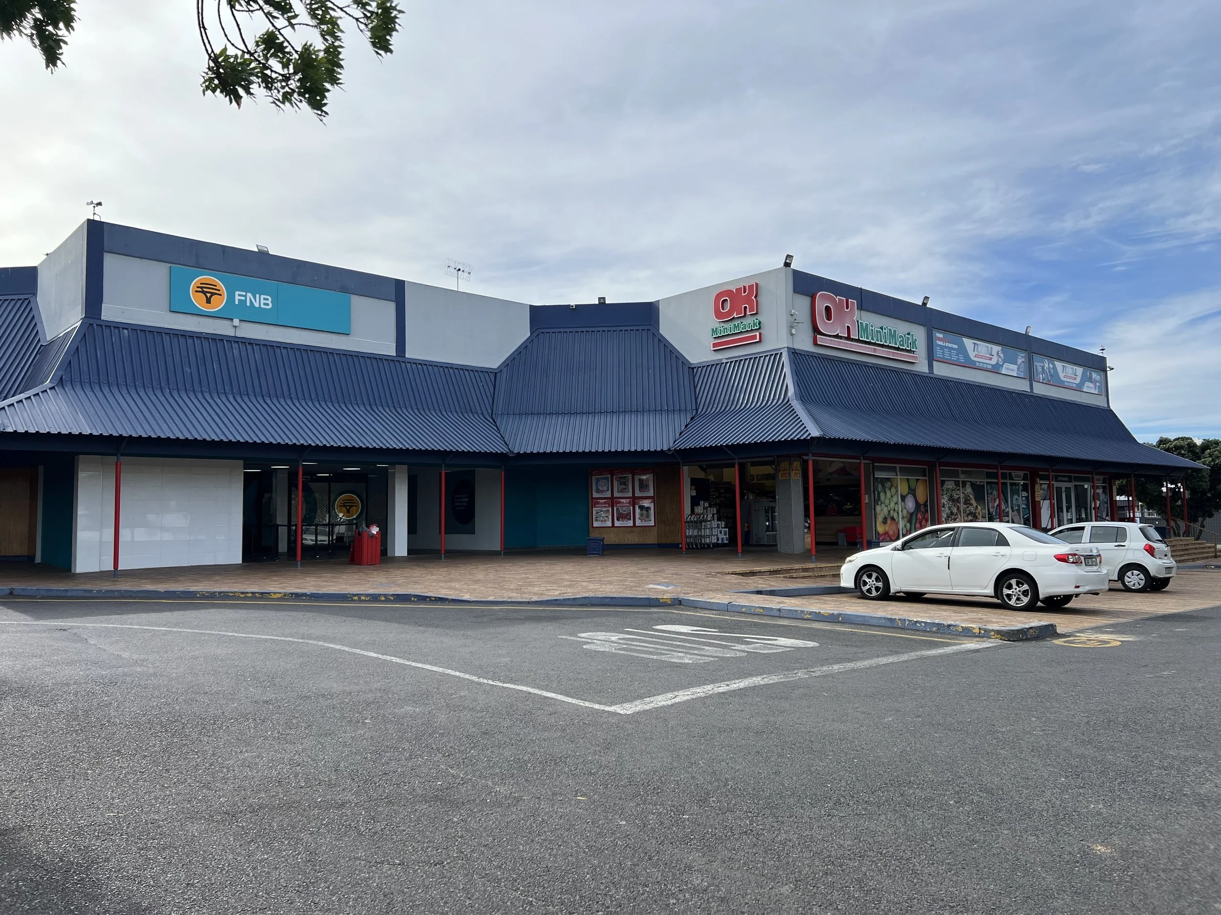 Shopping center with FNB bank and OK Multipharms signs, blue and white exterior, parked cars.
