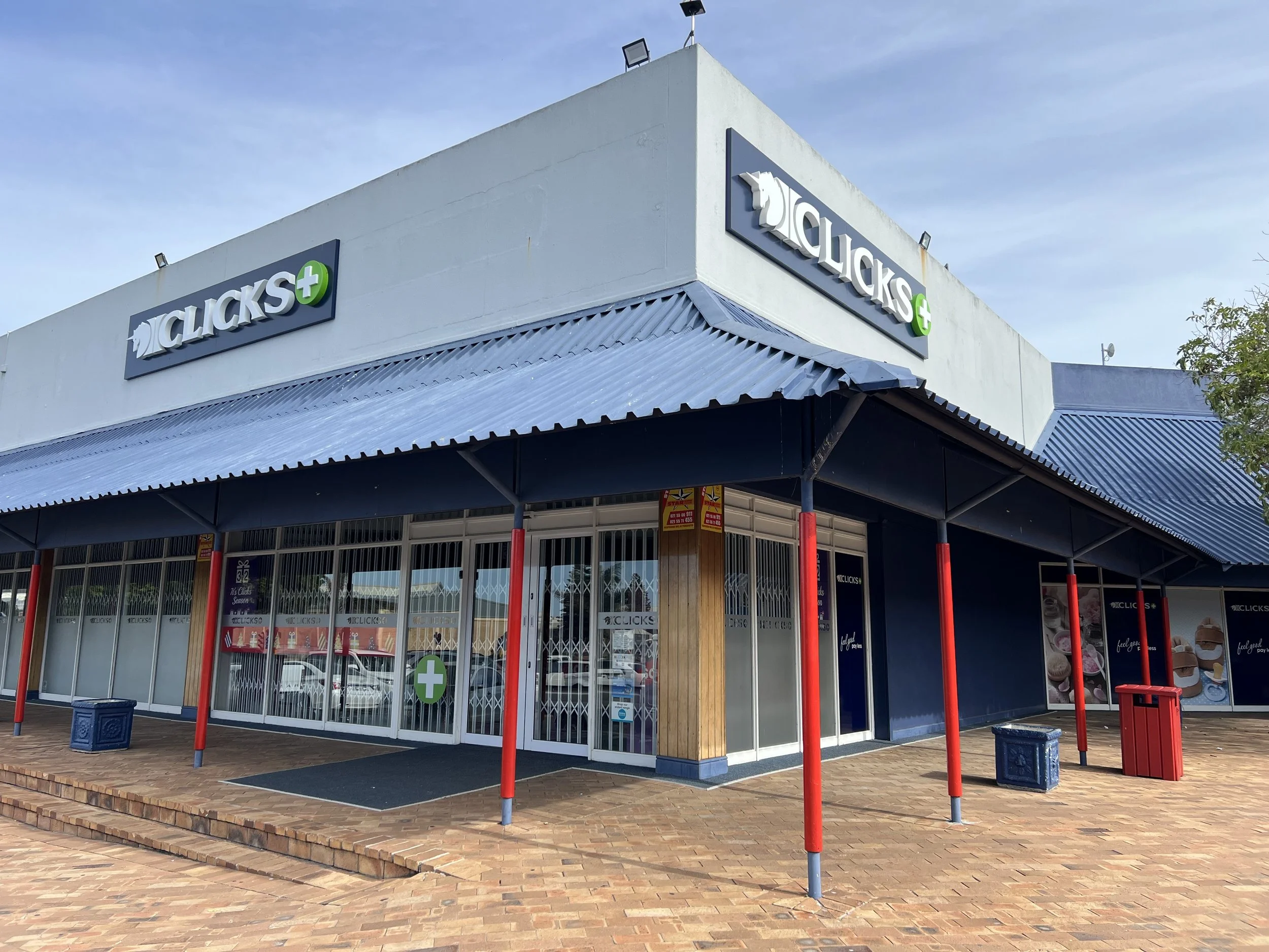 Exterior of a Clicks store with blue awning and red columns, featuring a green cross symbol at the entrance.