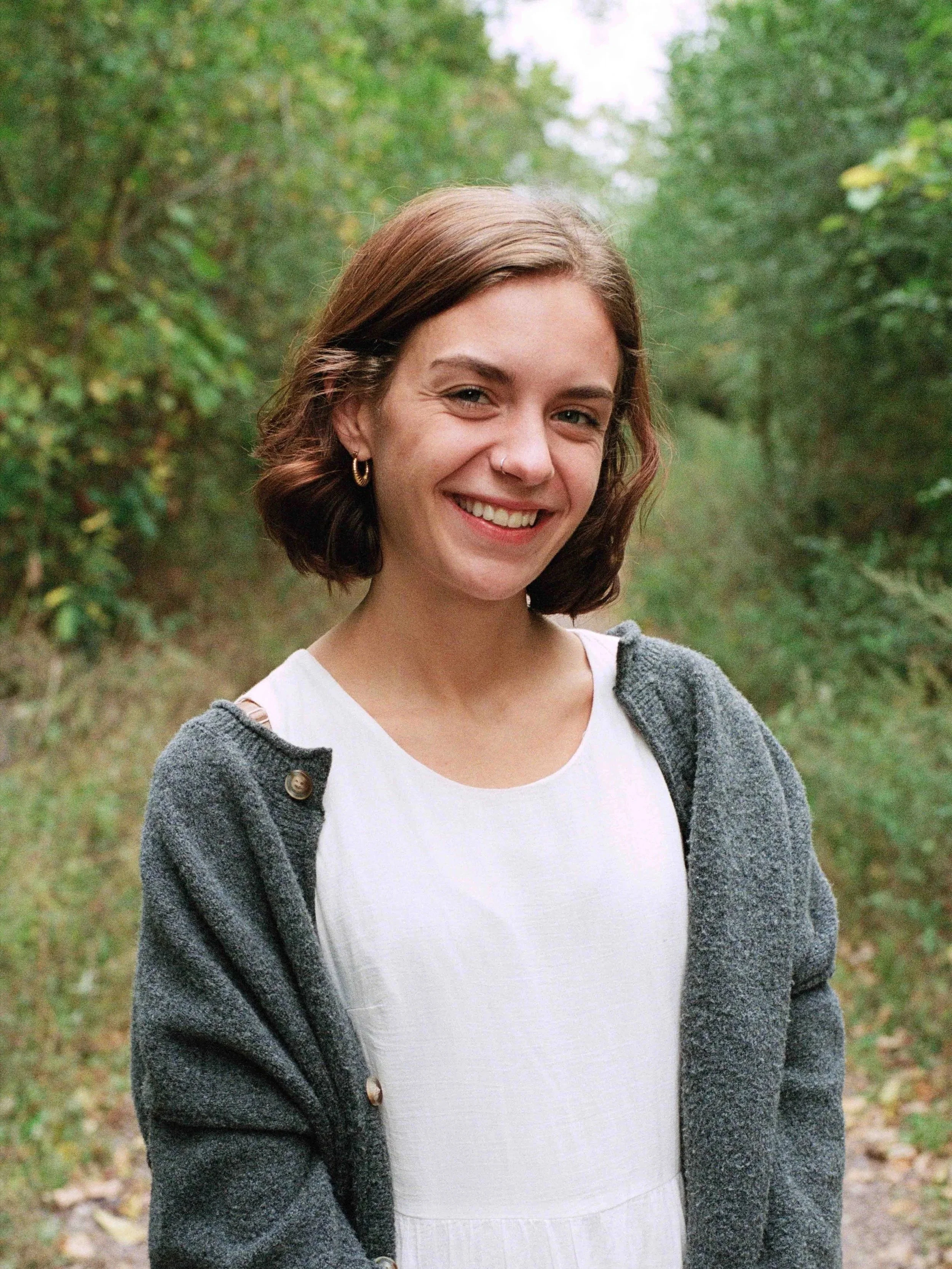 A young woman with short brown hair smiling outdoors in a wooded area, wearing a gray jacket over a white top.