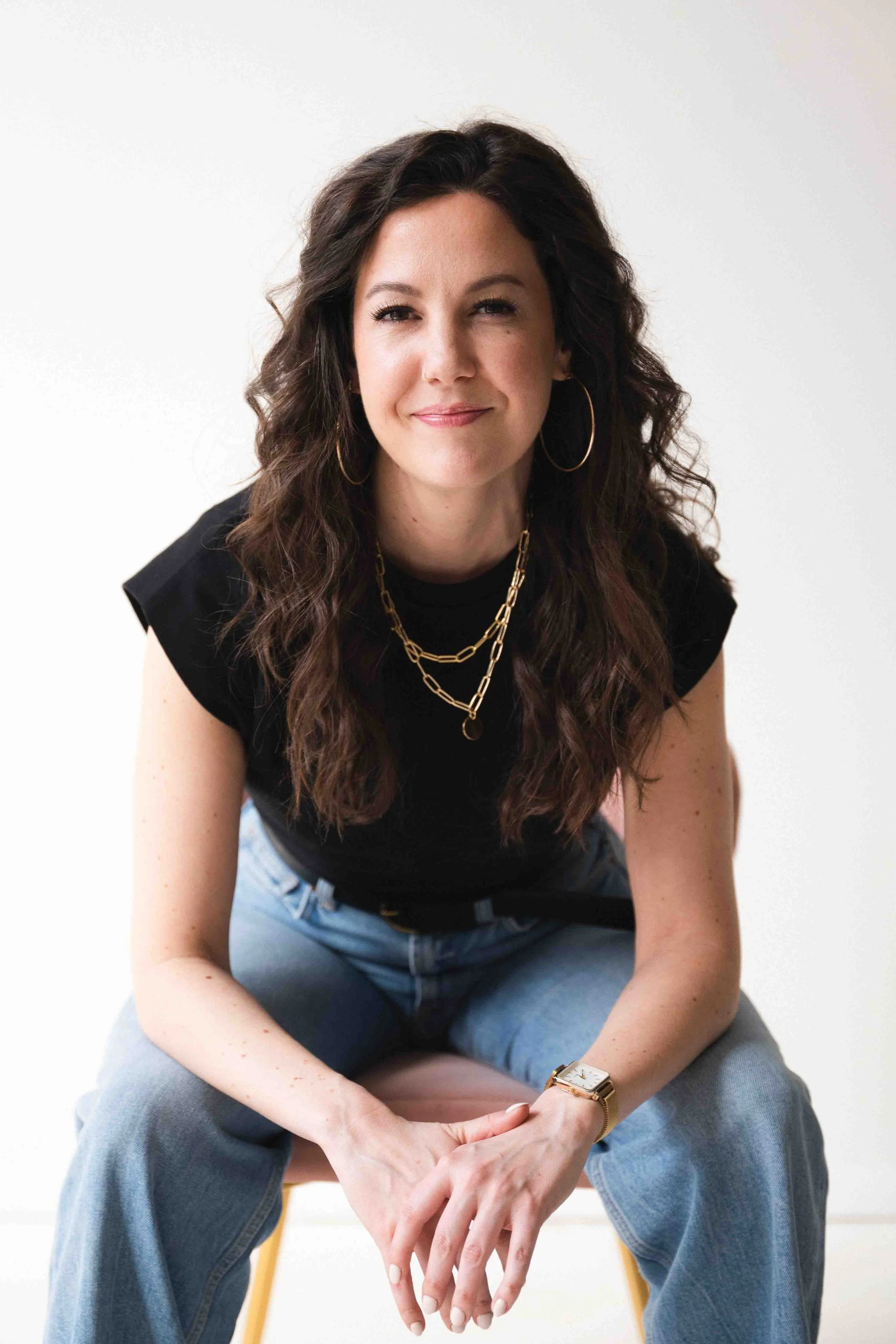 A woman with long curly brown hair, wearing a black top, gold hoop earrings, layered gold necklaces, and a gold wristwatch, sitting on a yellow chair against a plain white background.