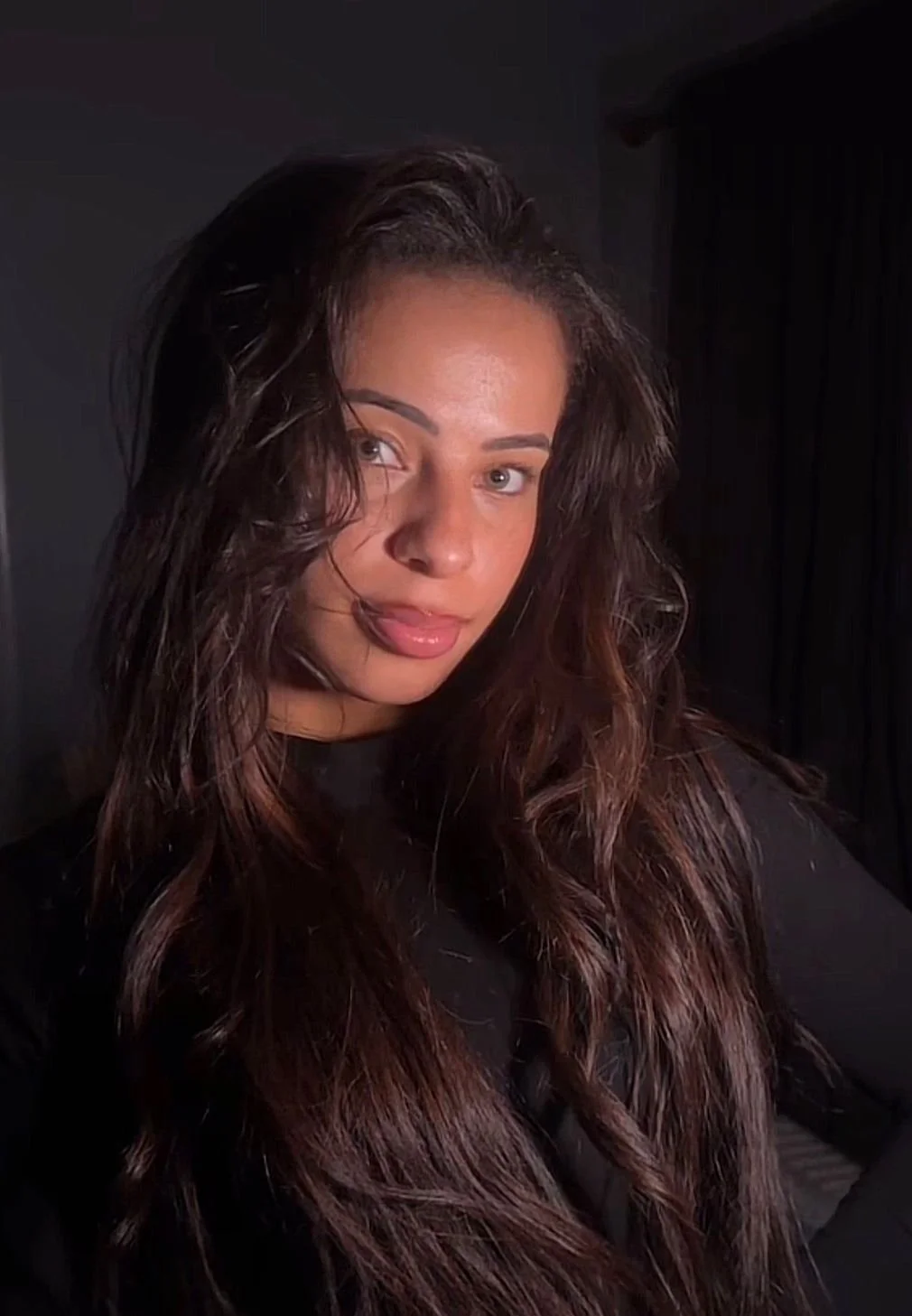 A woman with long, curly, brown hair and light skin looking at the camera with her tongue slightly out, in a dark room.