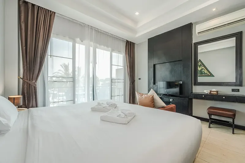 A modern hotel room with a large bed, white linens, folded towels, and a window with sheer curtains. There is a sitting area with an orange armchair, a flat-screen TV, a mirror, and a desk with a bench.