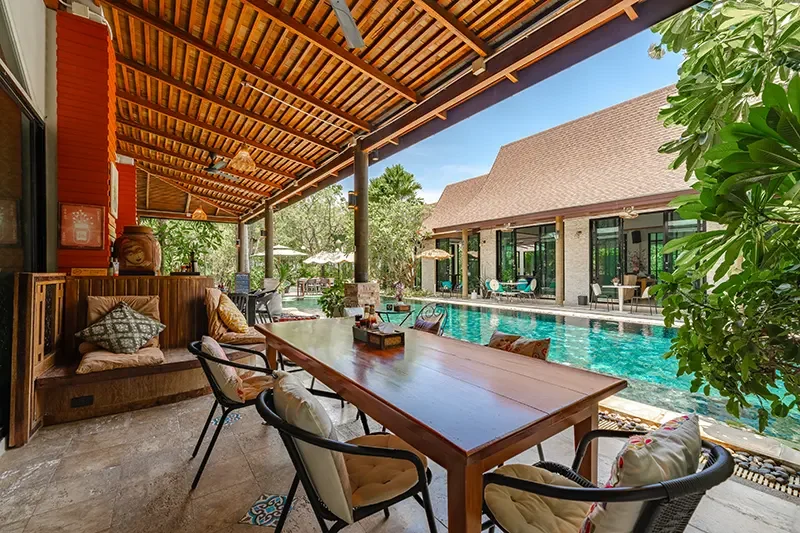 Outdoor patio with a wooden dining table and chairs, overlooking a swimming pool surrounded by lounge chairs, umbrellas, and a covered sitting area attached to a house.