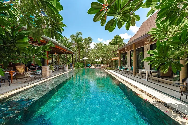 A luxurious outdoor swimming pool surrounded by lounge chairs, umbrellas, and tropical greenery, with a clear blue sky overhead.