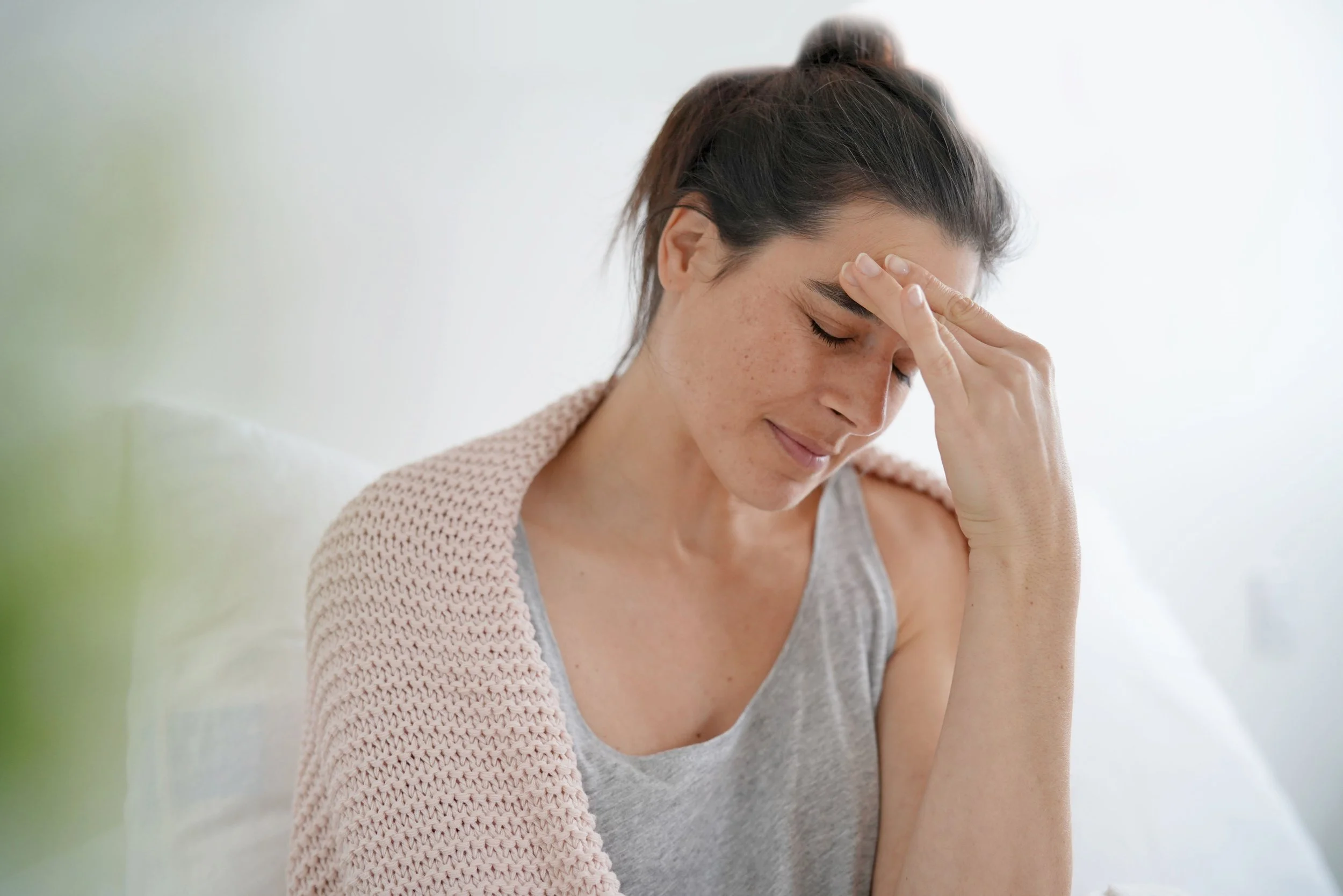 A woman with dark hair, wearing a gray tank top, sitting on a white couch, gently touching her forehead with a tired expression.